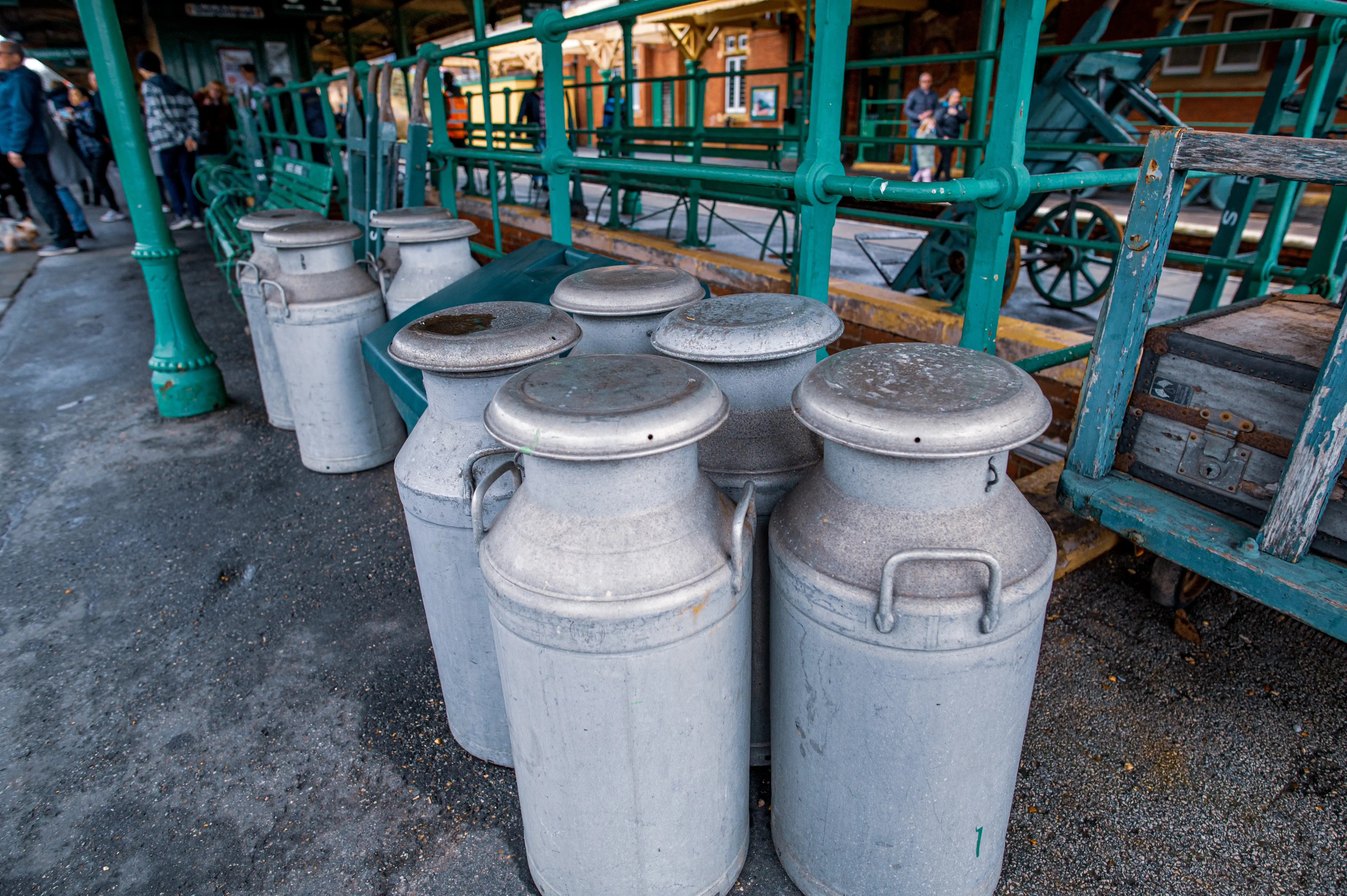 Row of vintage aluminium milk churns with metal handles and domed lids, some with water residue, lined up on a gravelly platform next to a green railing at a railway station. A blue wooden cart is visible to the right.