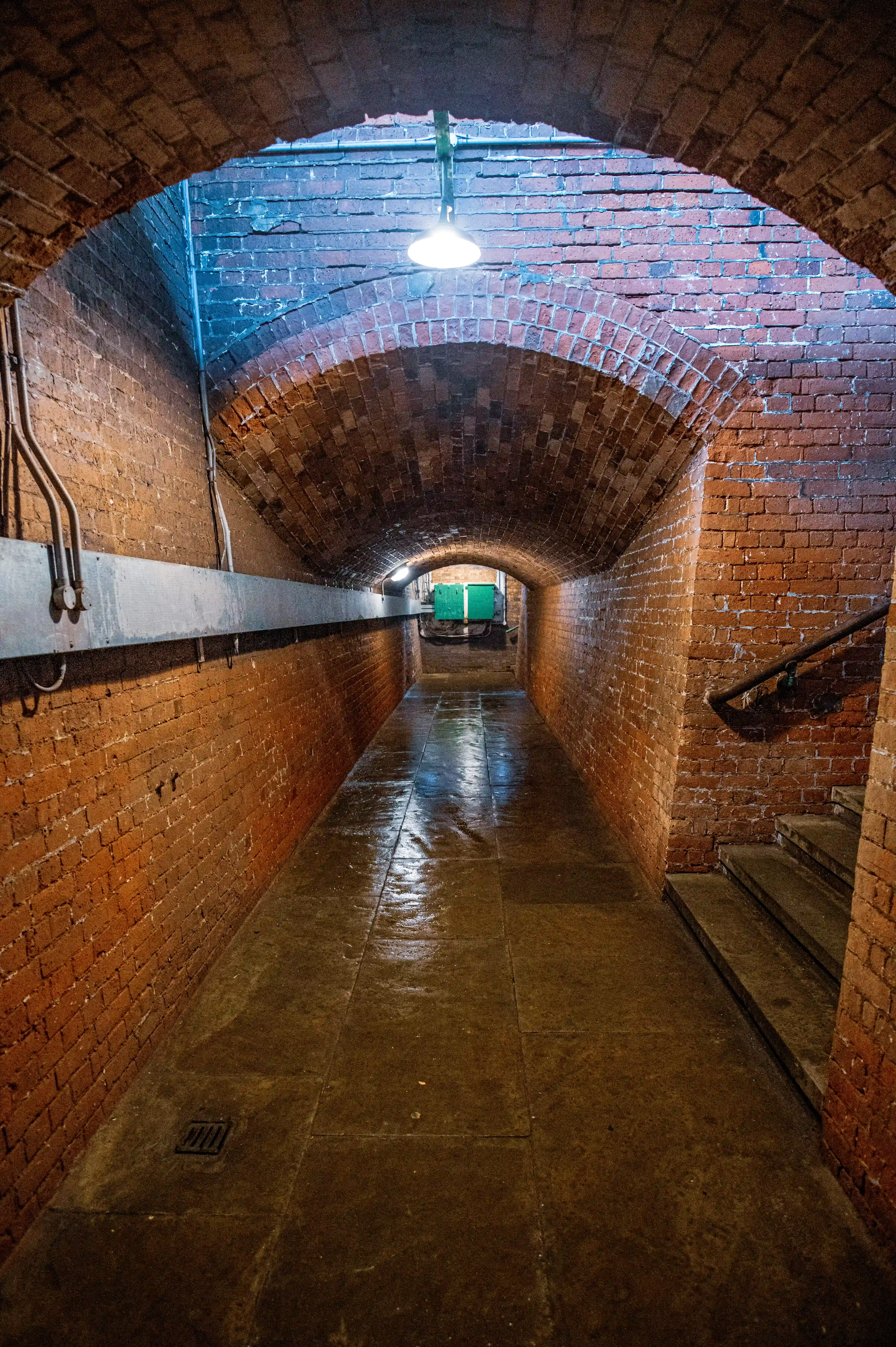 A long, narrow, arched brick tunnel with a damp, reflective floor. A single light hangs from the ceiling, illuminating the red brickwork and a set of steps on the right. Green doors are visible at the far end of the tunnel.