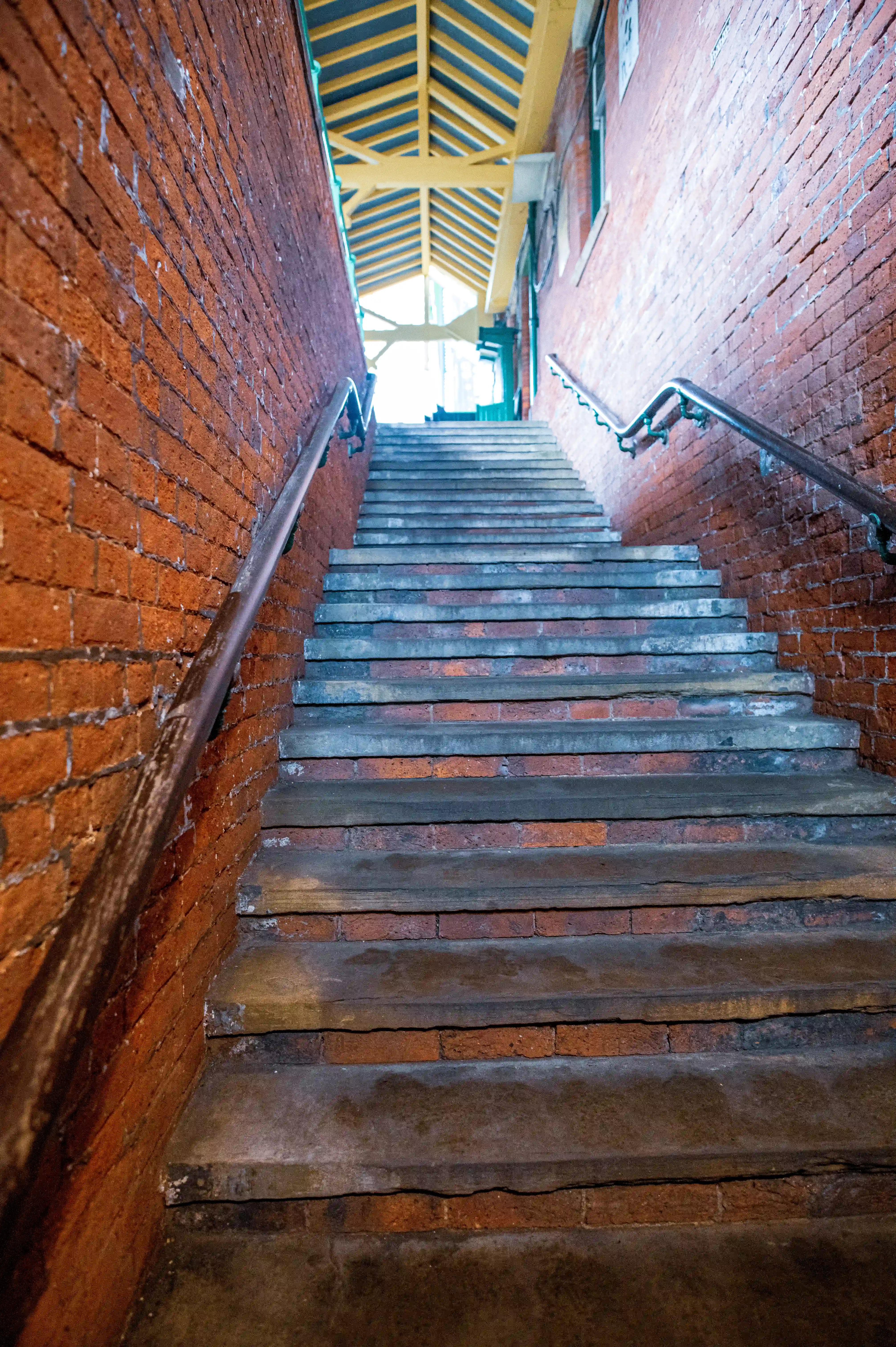 An outdoor perspective view looking up a set of stone and brick steps, bordered by rough red brick walls. A silver handrail runs along the right side. The stairway leads to a bright, open area with a yellow-roofed structure.