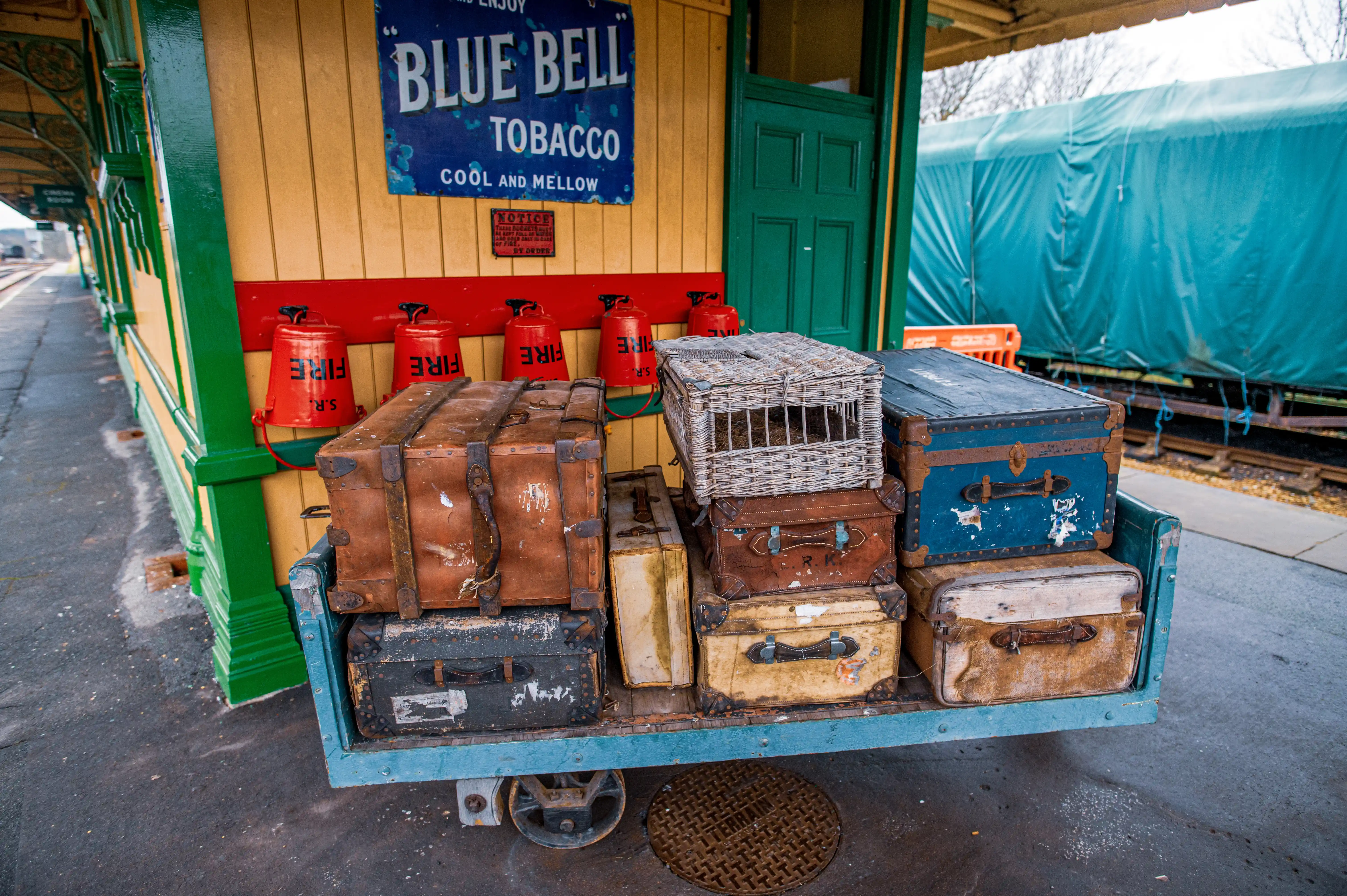 A close-up view of a trolley laden with vintage luggage, including several leather and wooden suitcases and a wicker pet carrier, parked on a railway platform. Red fire buckets and a vintage tobacco advertisement are visible on the yellow-painted station wall.