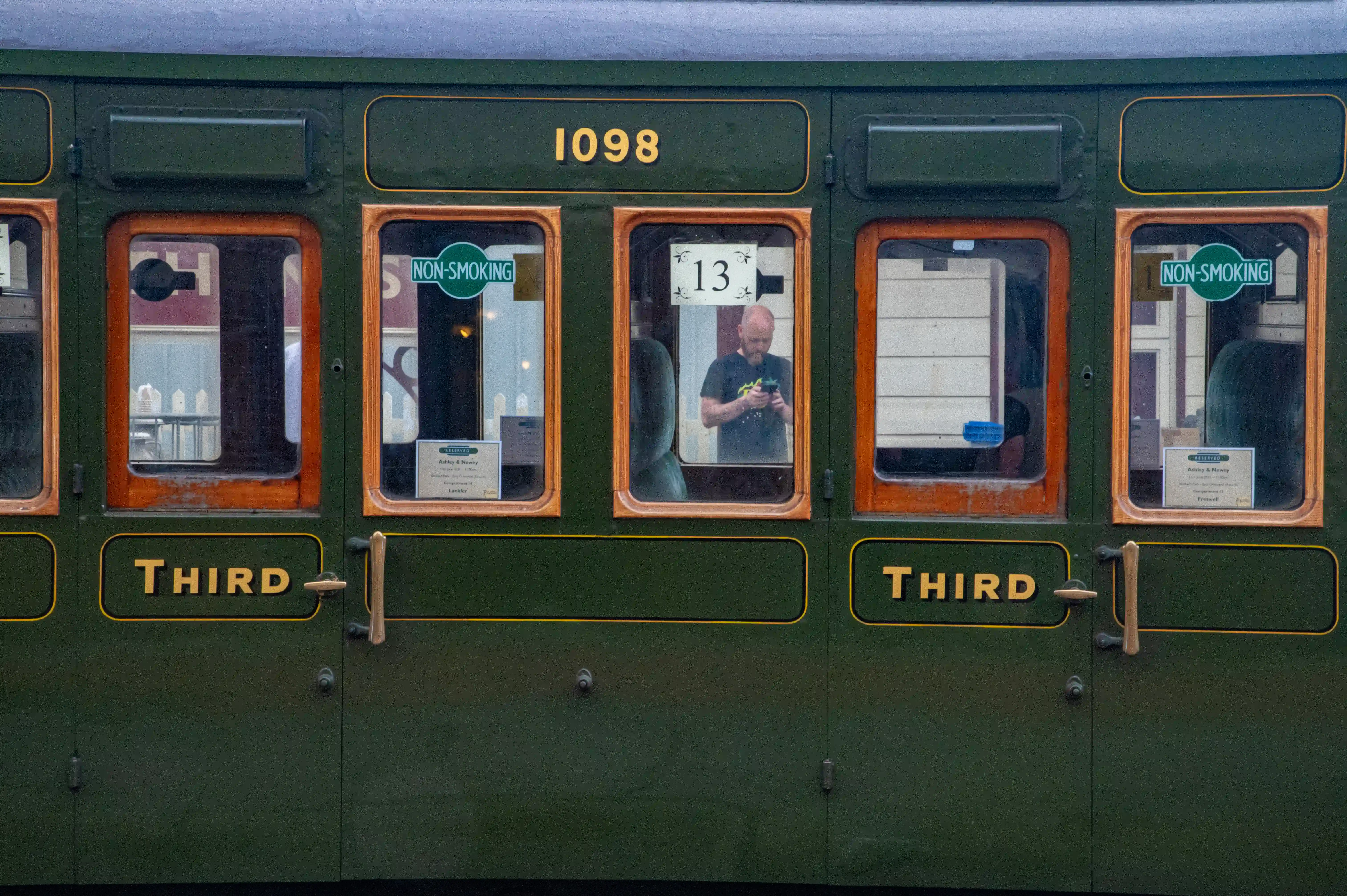 Exterior of a dark green vintage train carriage with wooden window frames. The number 1098 is visible above the windows, and 'THIRD' is written in yellow on the carriage body. A man is seen through one of the windows, looking at his phone, and his reflection is visible in the glass.