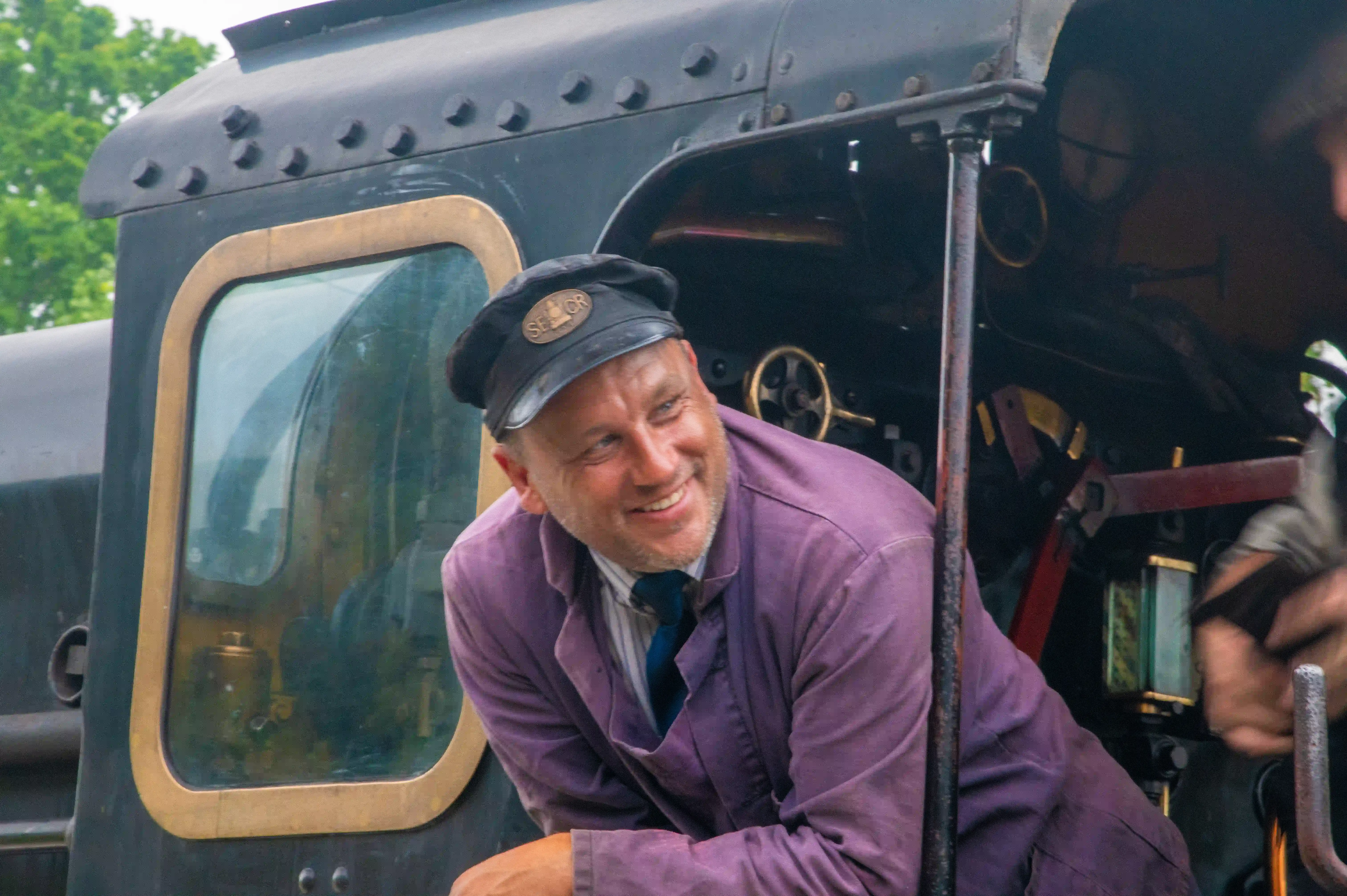 A middle-aged man with a beard and a peaked cap with a 'SECTOR' badge is looking to his right with a wide, happy smile. He is wearing a striped shirt, a dark blue tie, and a purple jacket. He is leaning out of the driver's cab of a black steam engine, with the yellow-trimmed window of the cab to his left and various mechanical parts of the engine visible behind him.