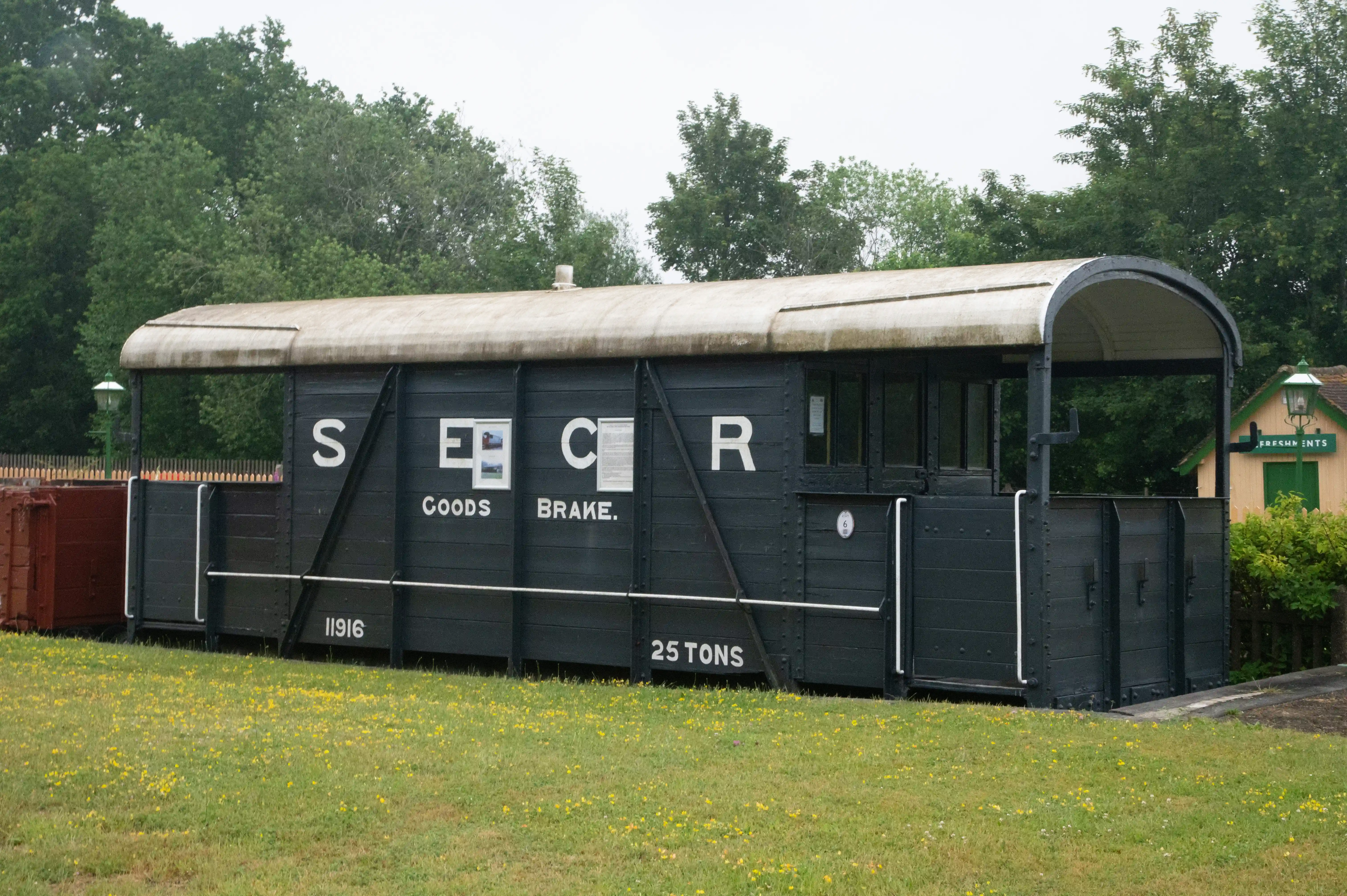 A dark grey, wooden railway wagon with a curved roof sits on tracks. White lettering on the side reads 