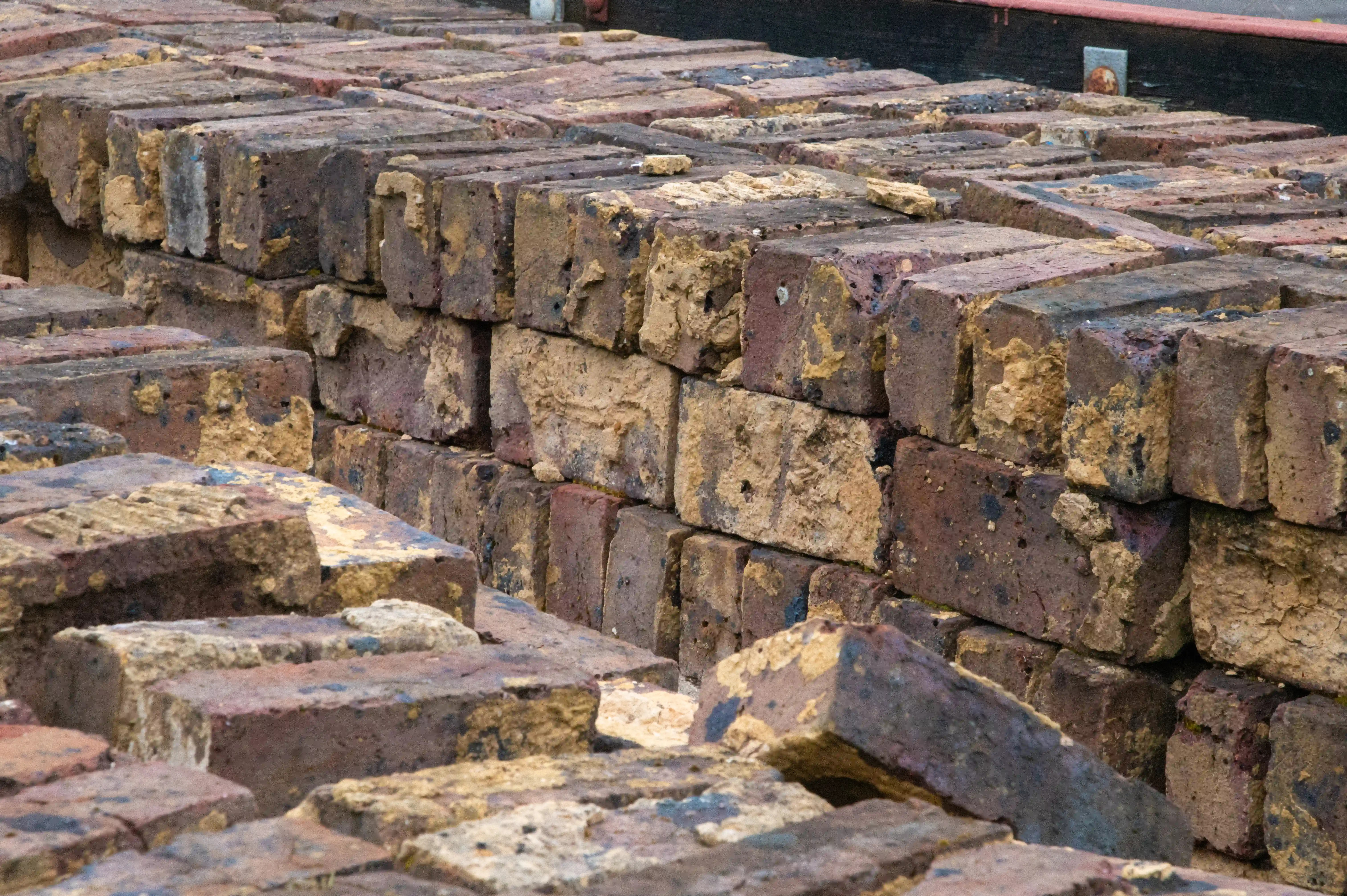 A textured close-up of a pile of old, weathered bricks, showing various shades of brown and yellow, with some chipped edges and rough surfaces.