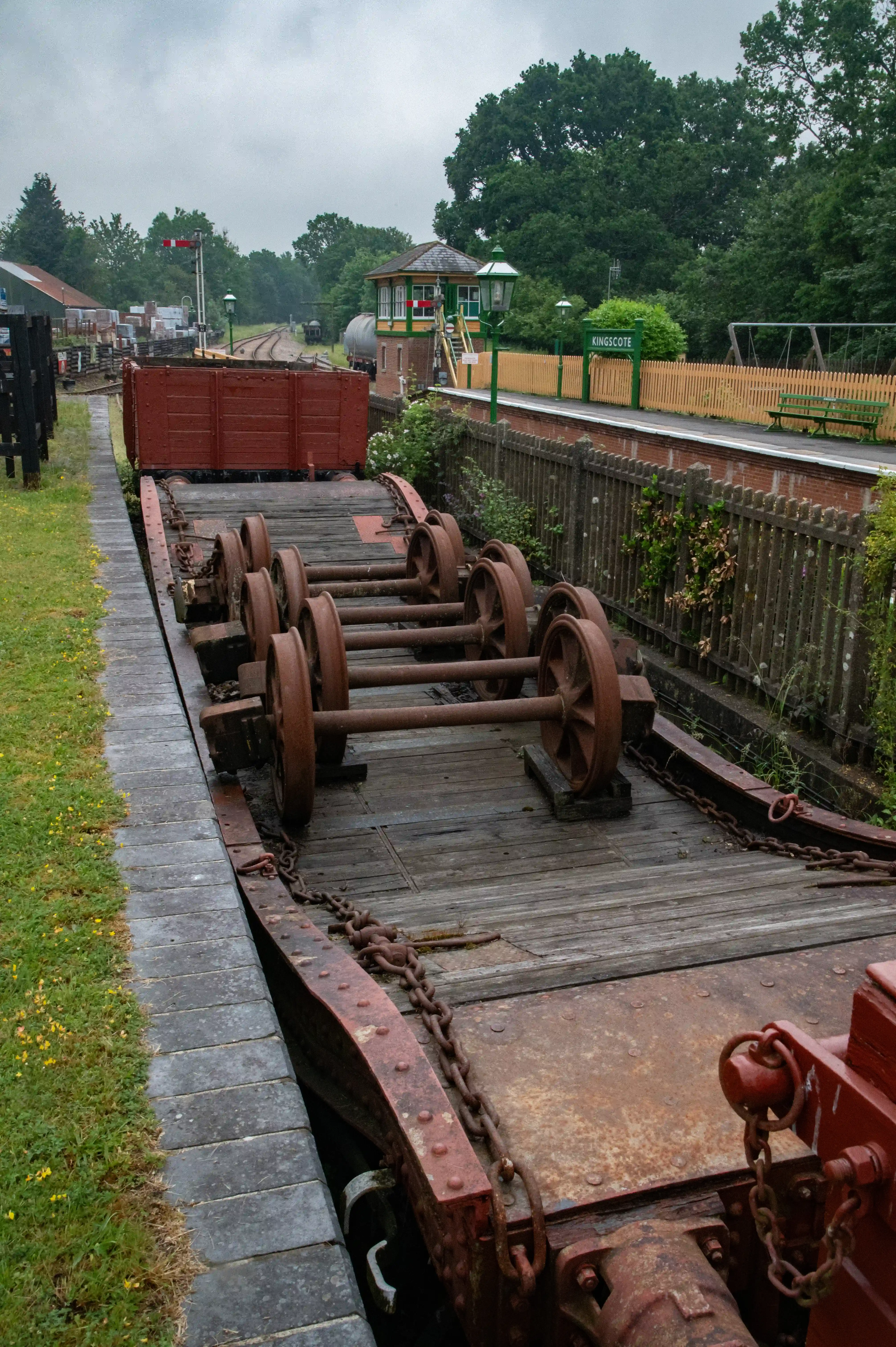 A rusty, red railway goods wagon with multiple sets of wheels is parked alongside a wooden platform at Kingscote station. A vintage signal box and signal are visible in the background under a cloudy sky, with trees surrounding the area.