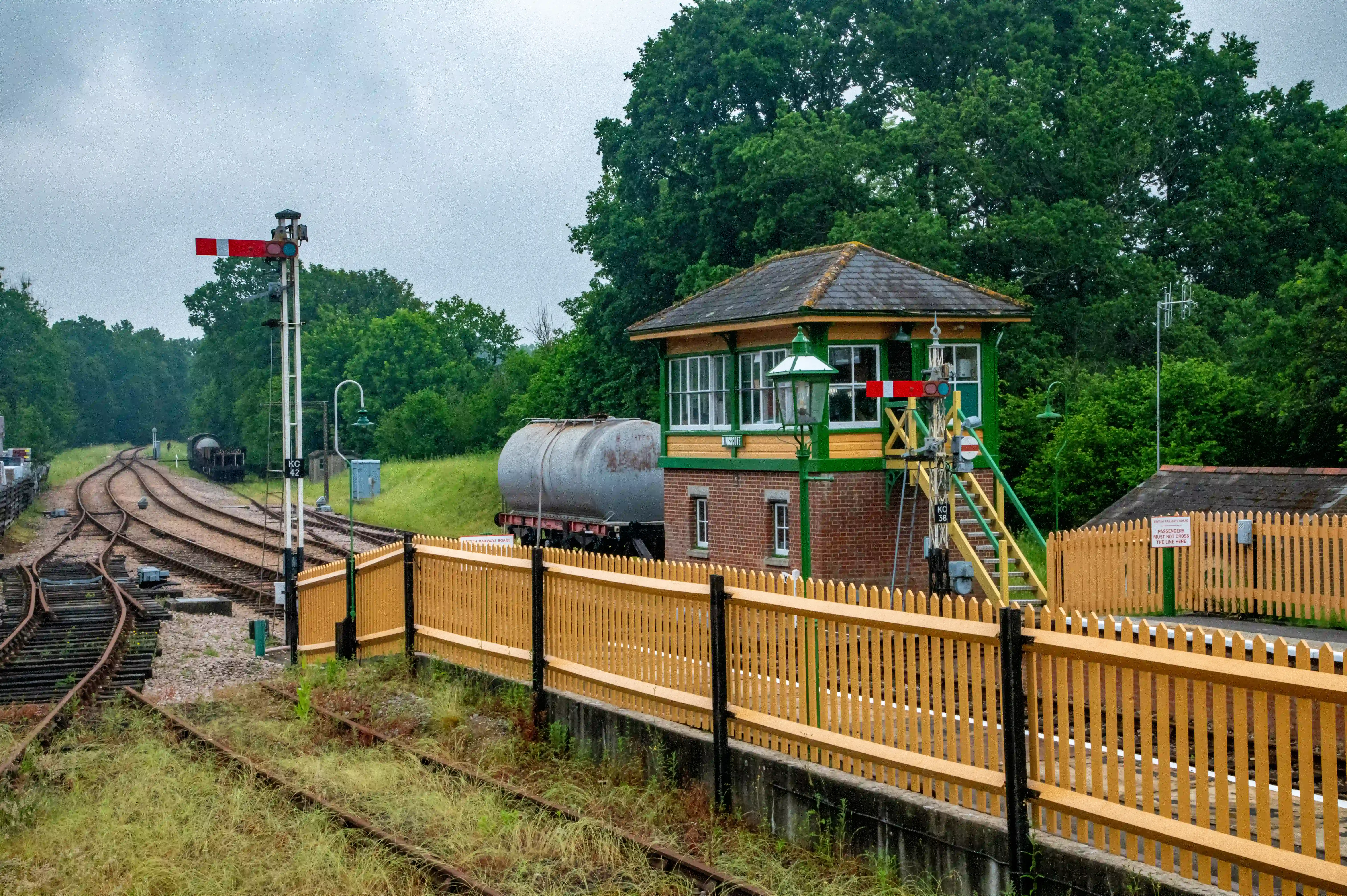 A railway signal box with green and yellow trim, next to a red and white semaphore signal, with railway tracks and a tanker wagon in the background.
