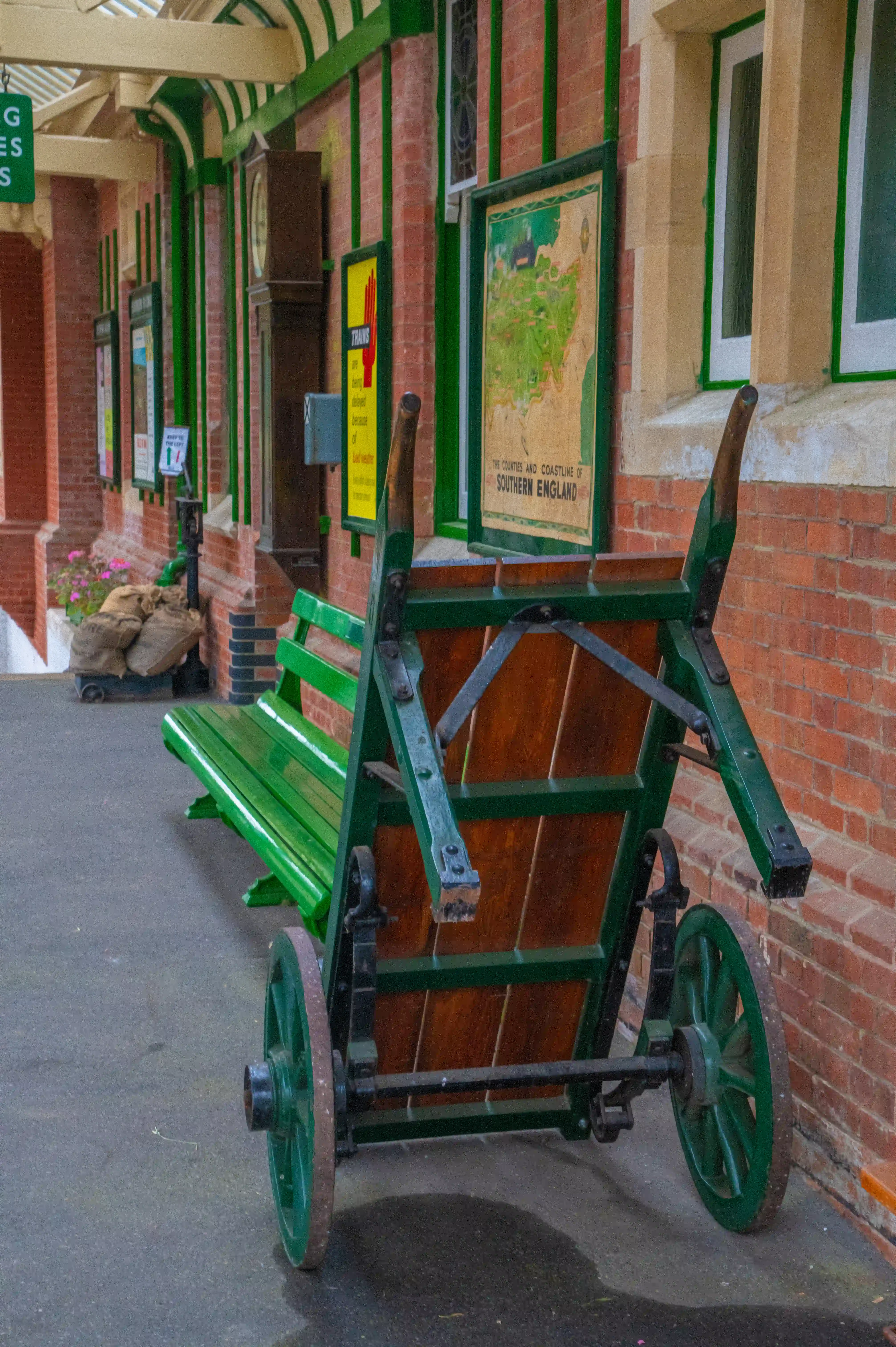 A green wooden handcart with large spoked wheels parked on a platform at a vintage railway station. The station has brick walls, green-painted wooden pillars, and vintage posters, including a map of 'The Counties and Coastline of Southern England.' A green bench and potted flowers are also visible