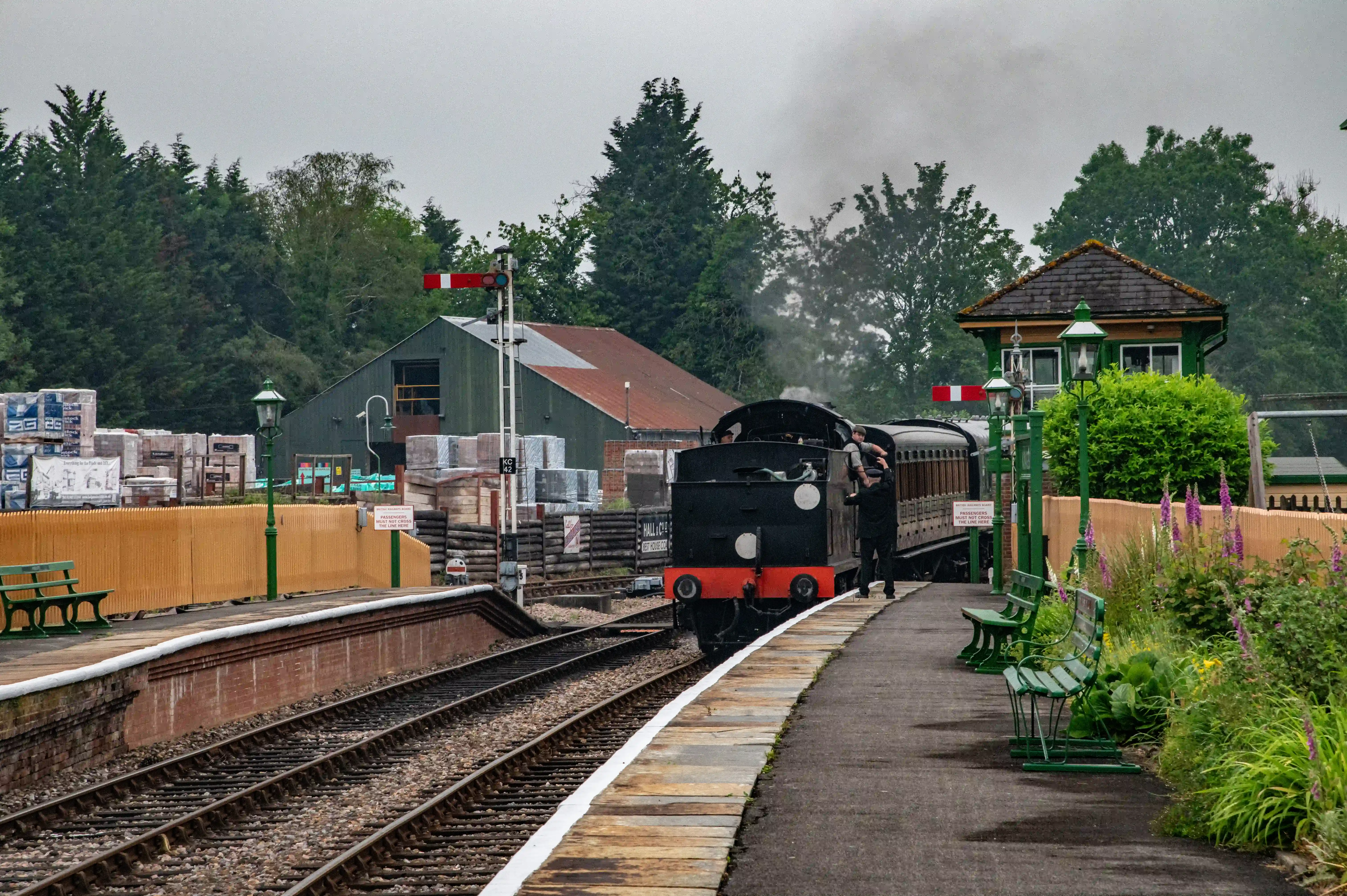 A black steam locomotive with red detailing is pulling a train of old-fashioned carriages along a railway track. Dark smoke is billowing from the chimney. The train is at a station platform with green benches and flowerbeds.