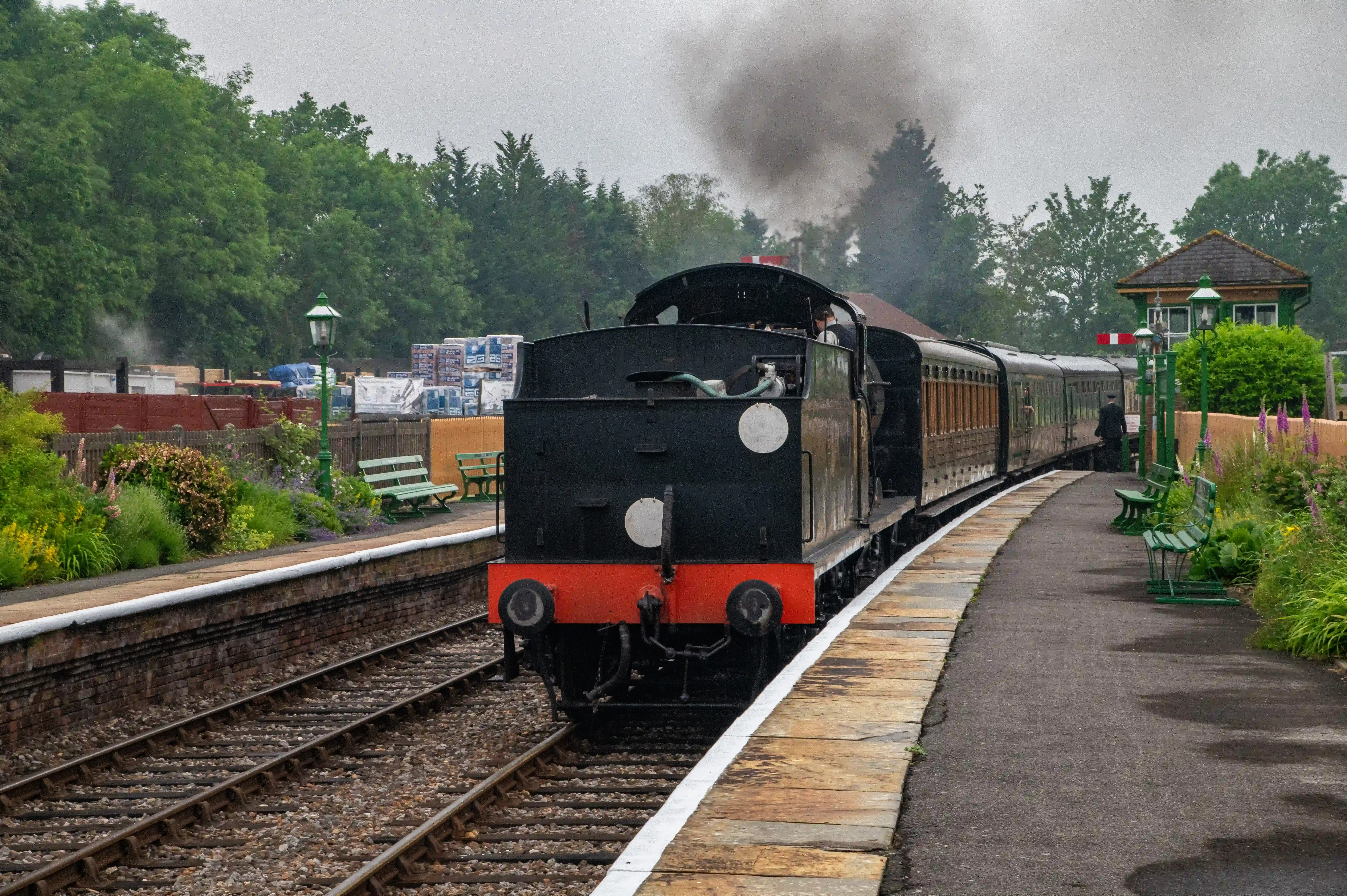 A black steam locomotive with red detailing is pulling a train of old-fashioned carriages along a railway track. Dark smoke is billowing from the chimney. The train is at a station platform with green benches and flowerbeds.