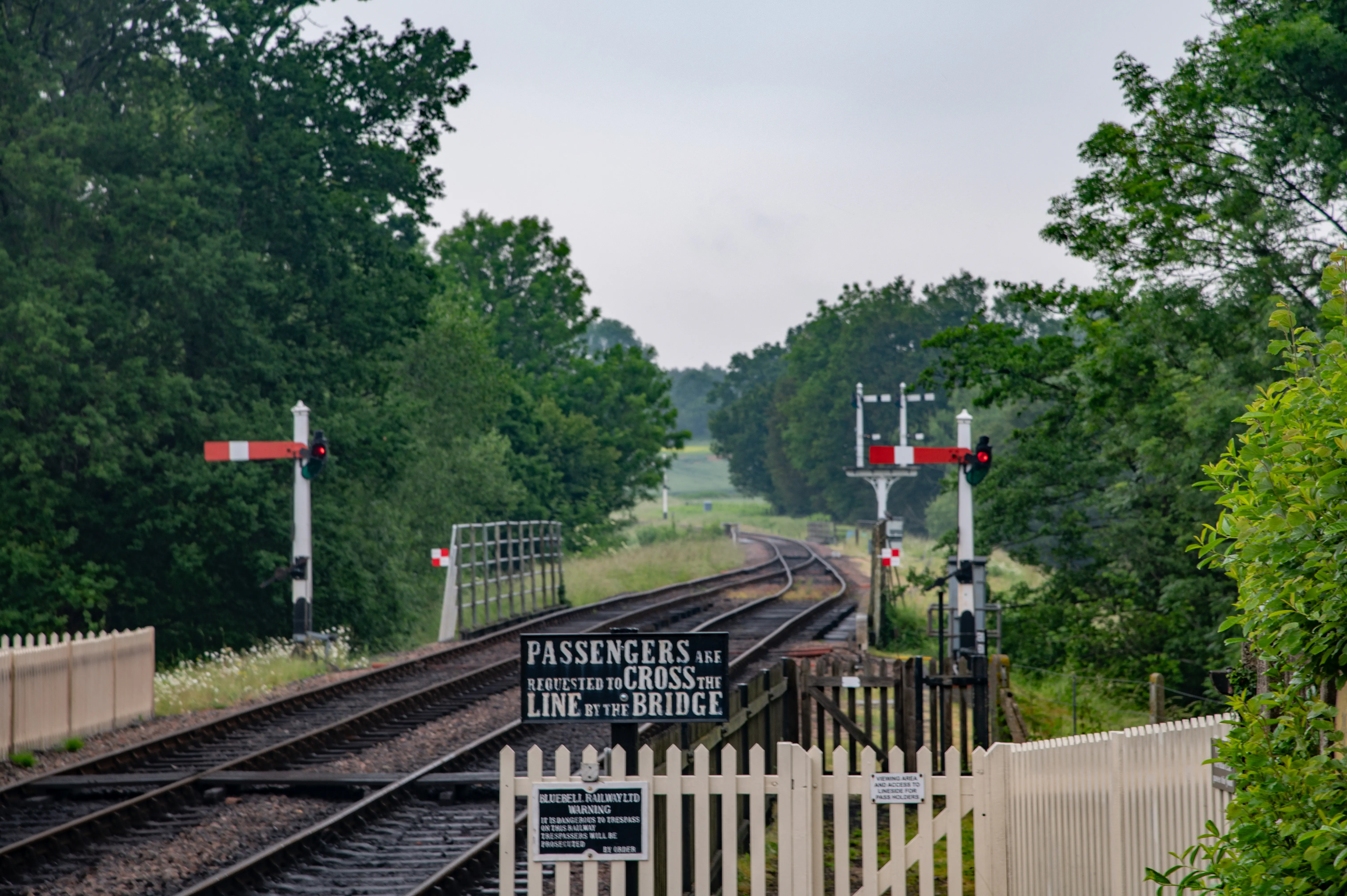 A country railway line with traditional red and white signals, passing through a wooded area. A sign reads PASSENGERS ARE REQUESTED TO CROSS THE LINE BY THE BRIDGE.