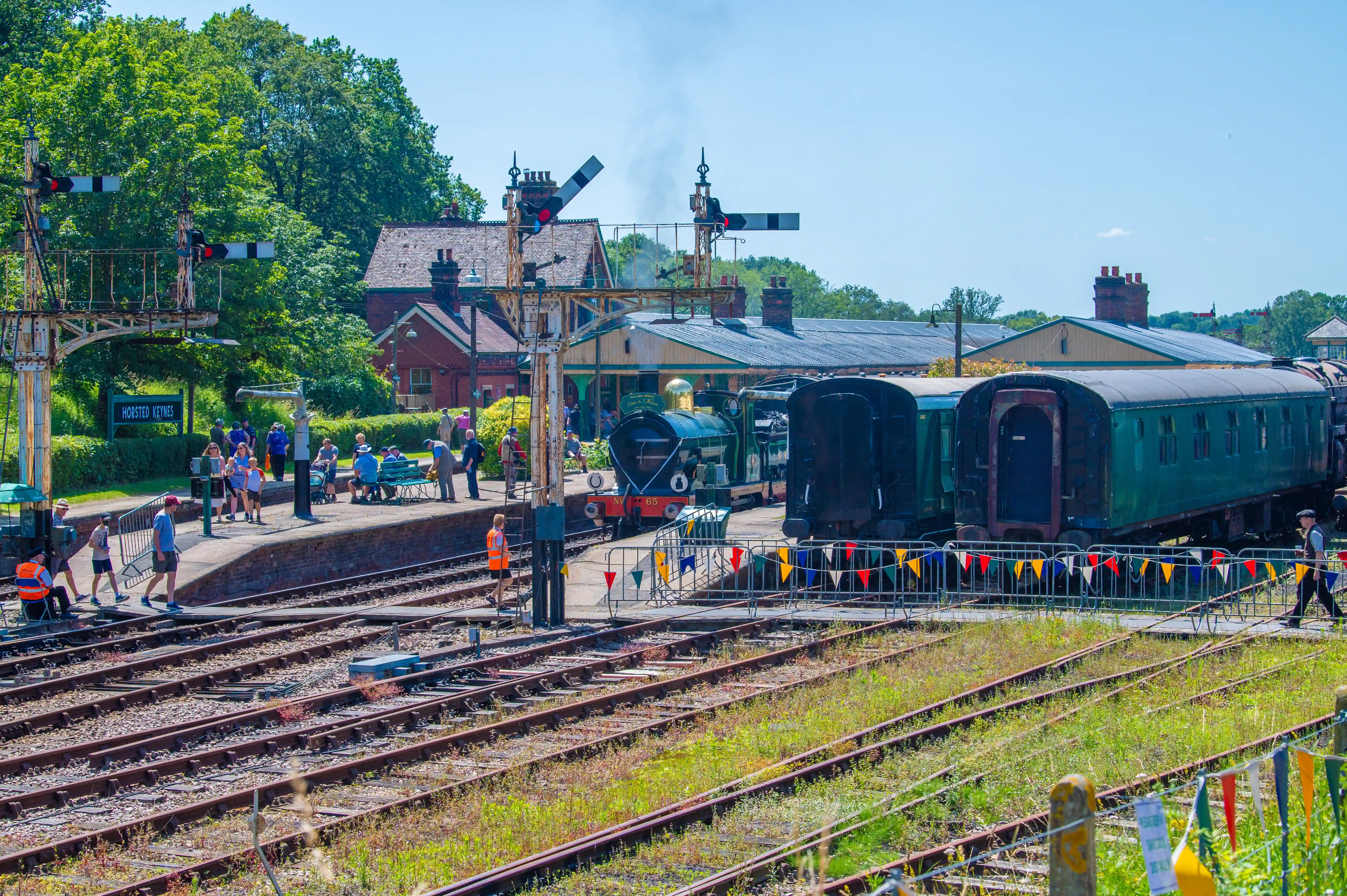 A steam train with carriages waits at a platform at the Horsted Keynes railway station, with people on the platform and signal posts visible.