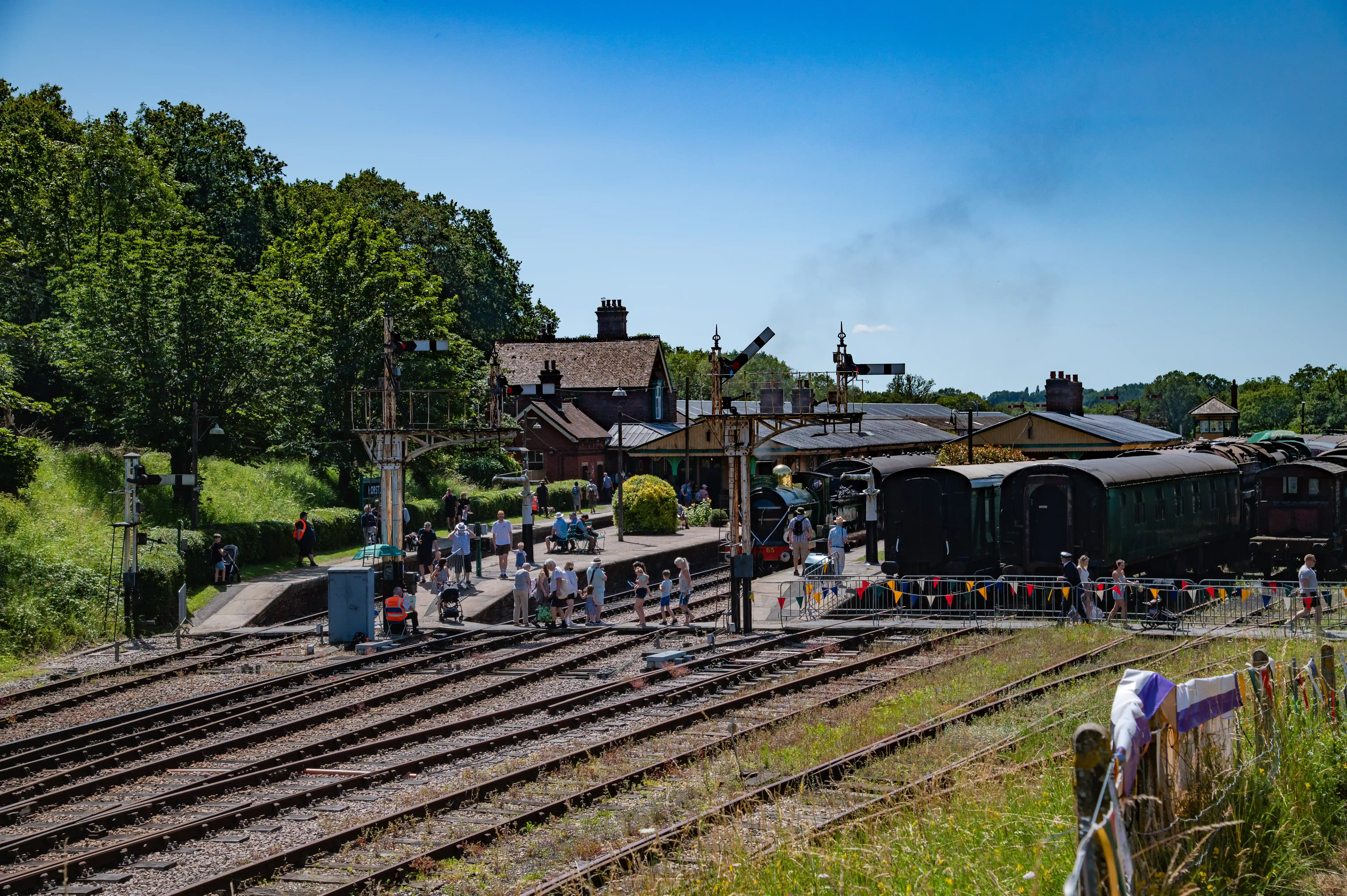 A broad view of a heritage railway station on a clear day. Multiple railway tracks run in the foreground, leading towards a platform where people are gathered. A steam train is visible with some carriages lined up. Traditional brick station buildings and railway signals are present, surrounded by lush green trees.