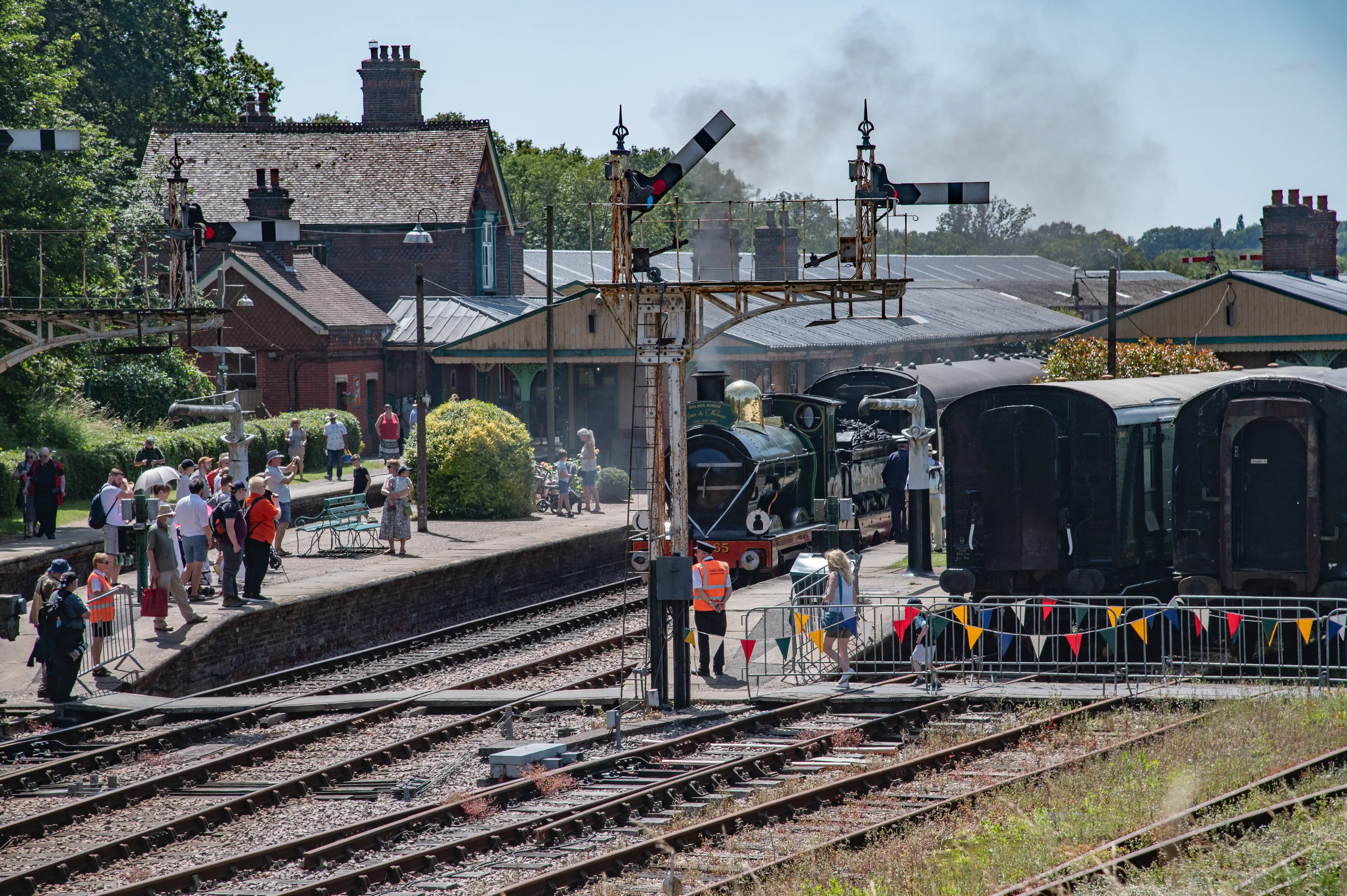 A vintage green steam train emitting smoke arrives at a historic brick railway station on a sunny day. People are gathered on the platform and along the tracks.
