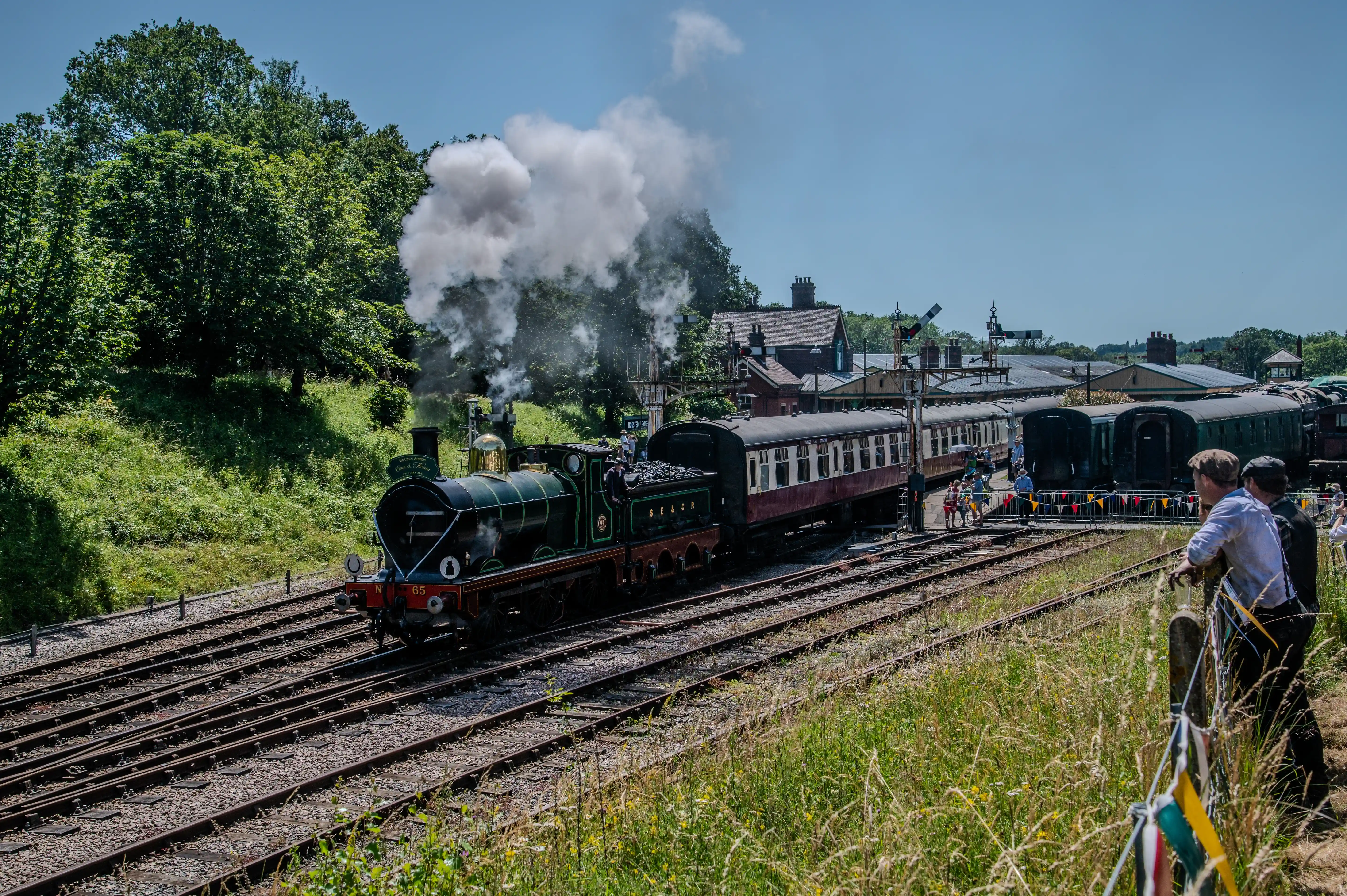 A green steam train numbered 65, with a plume of white smoke rising from its chimney, approaches a station with several carriages. People are visible on the platform and two men are looking towards the train from the right foreground.