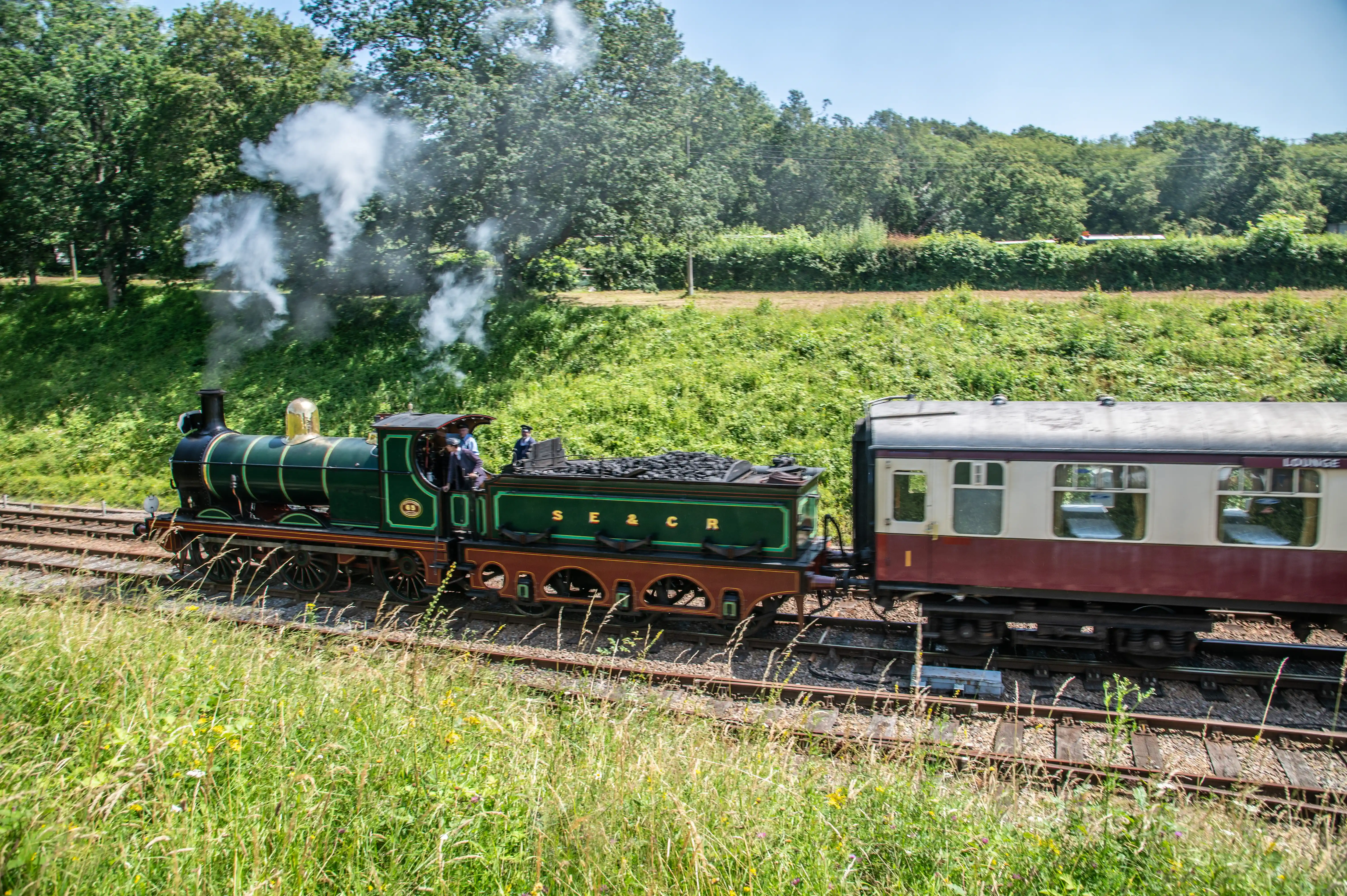 A vintage green steam train with the letters SE&CR on its tender chugs along a track, pulling a passenger carriage labelled 