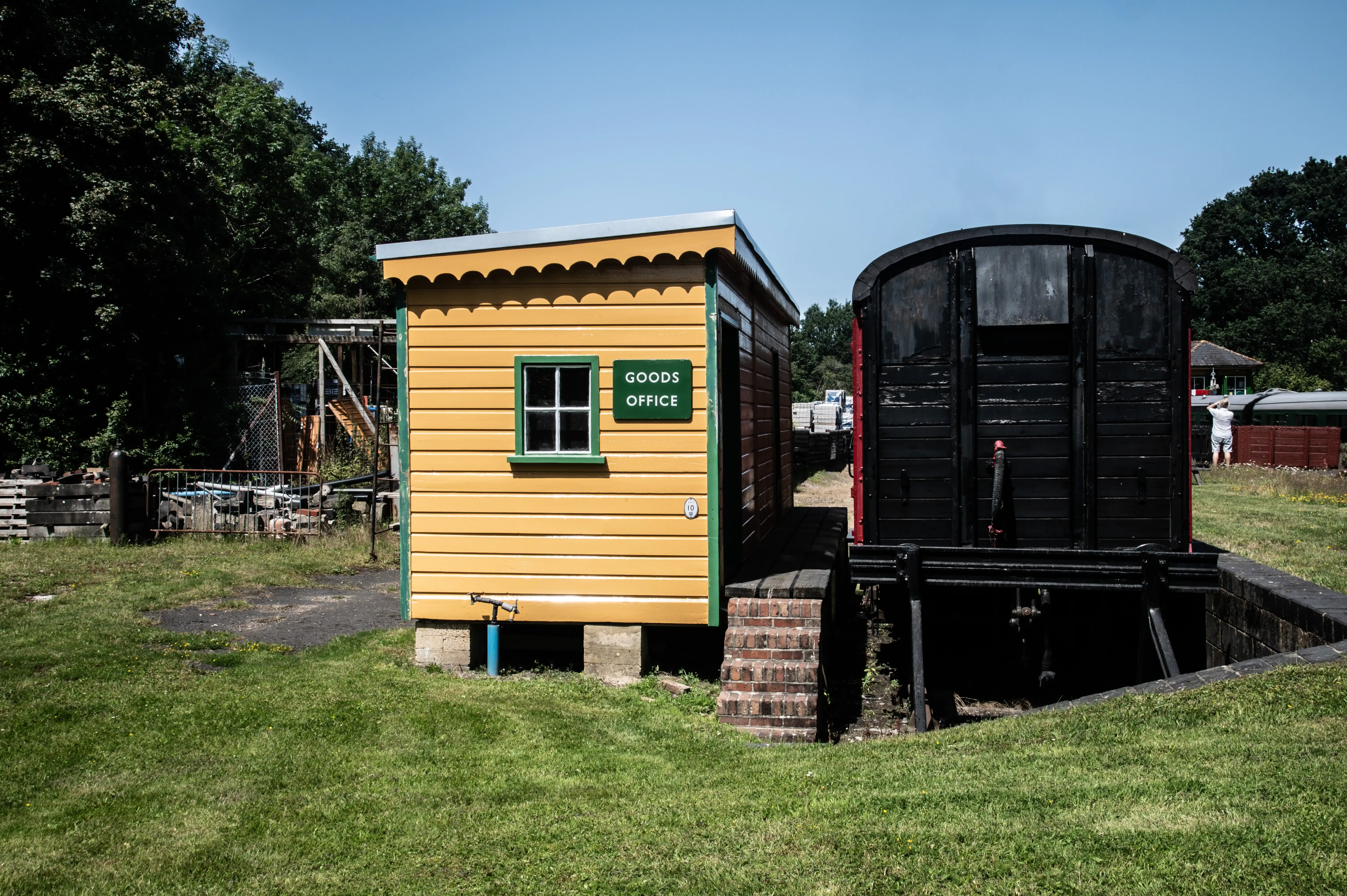 A small yellow wooden building with a green window frame and a sign reading GOODS OFFICE is positioned next to the side of a dark, older model train carriage. The scene is set outdoors on a bright, clear day with lush green grass and trees in the background.