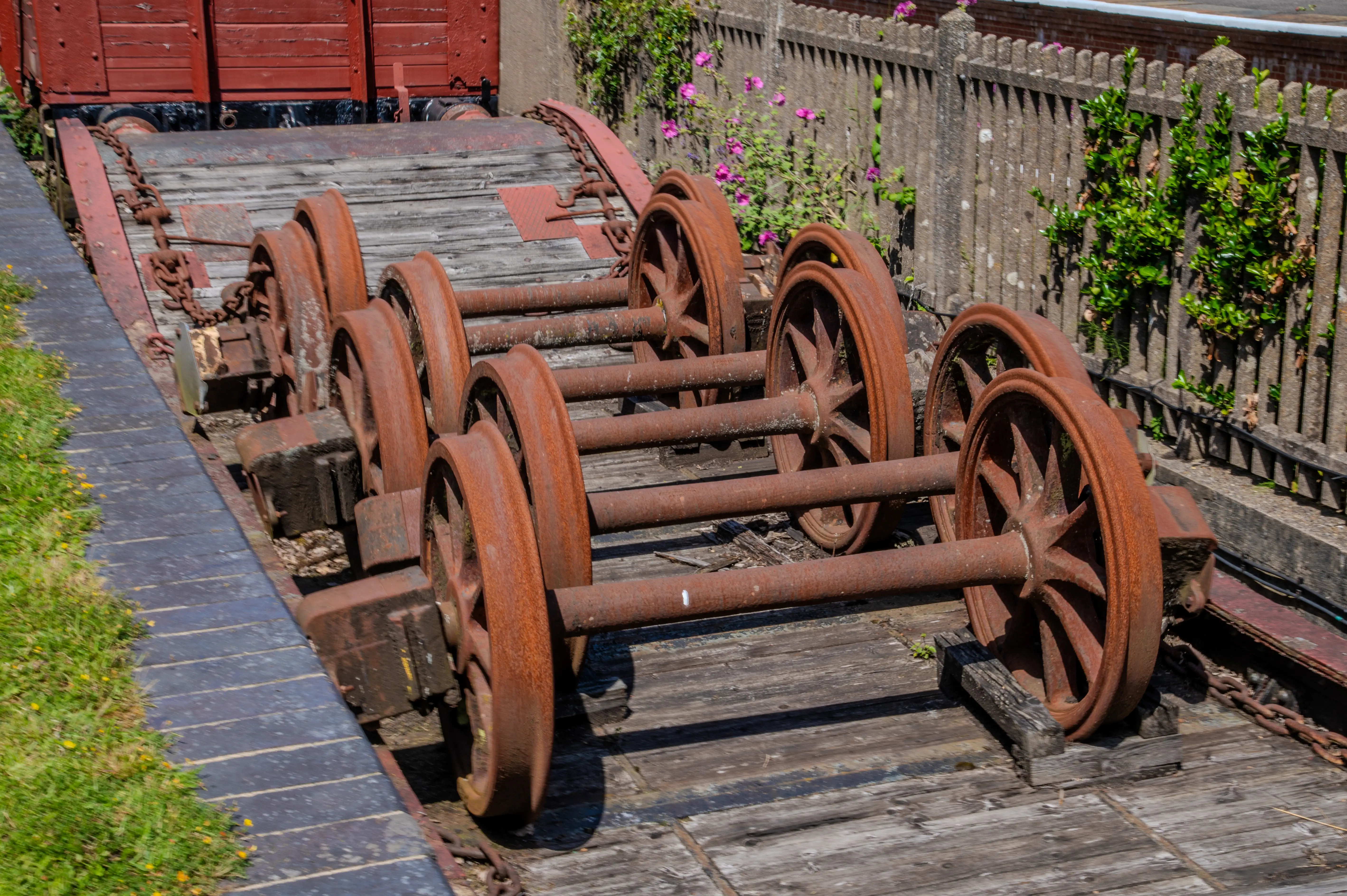 Several large, rusty, spoked railway wheels are positioned on a wooden platform. A red train car is visible in the upper left corner.