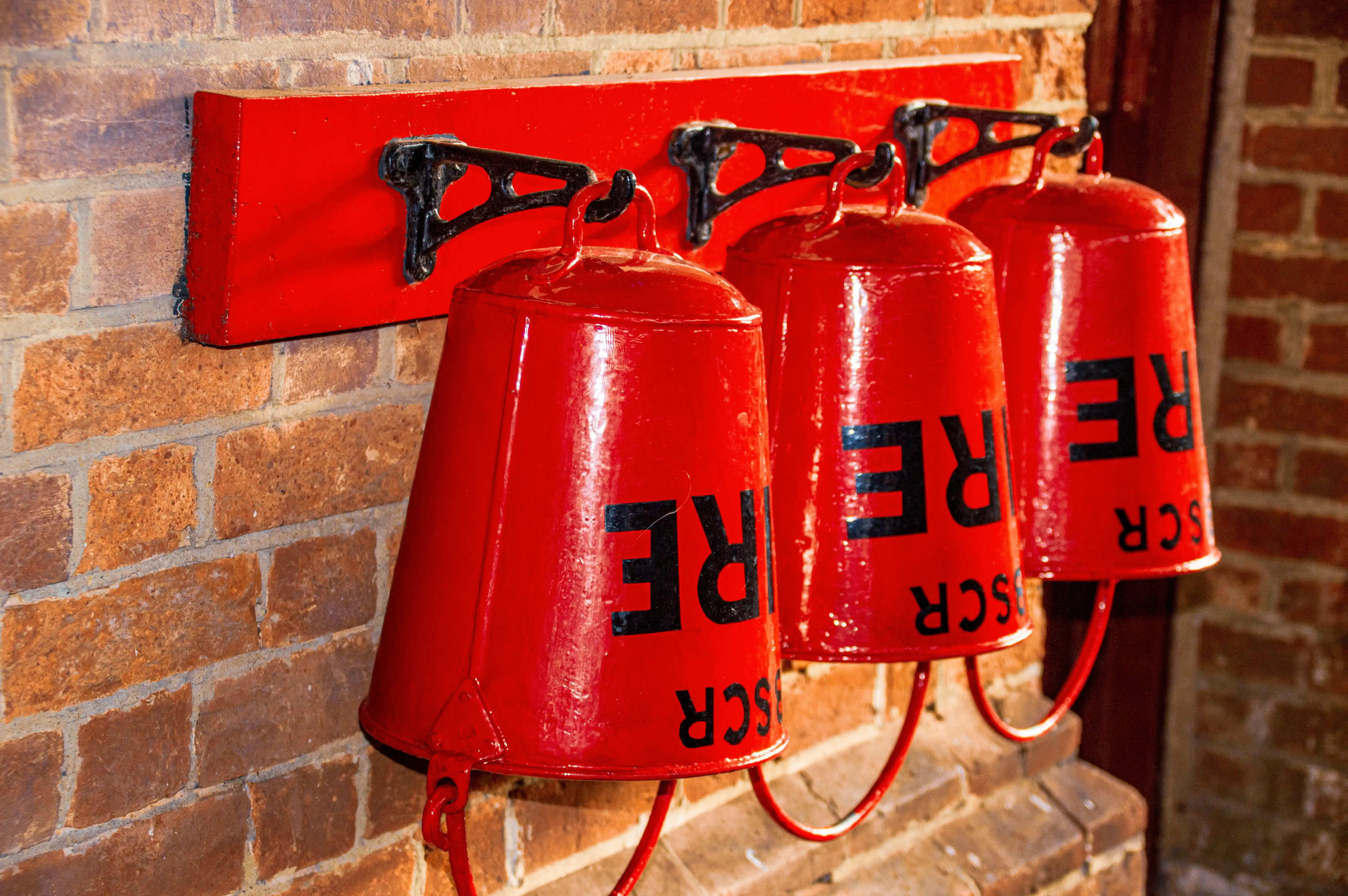 Three red fire buckets with 'FIRE' written on them in black letters, mounted on a red wooden plank attached to a brick wall.