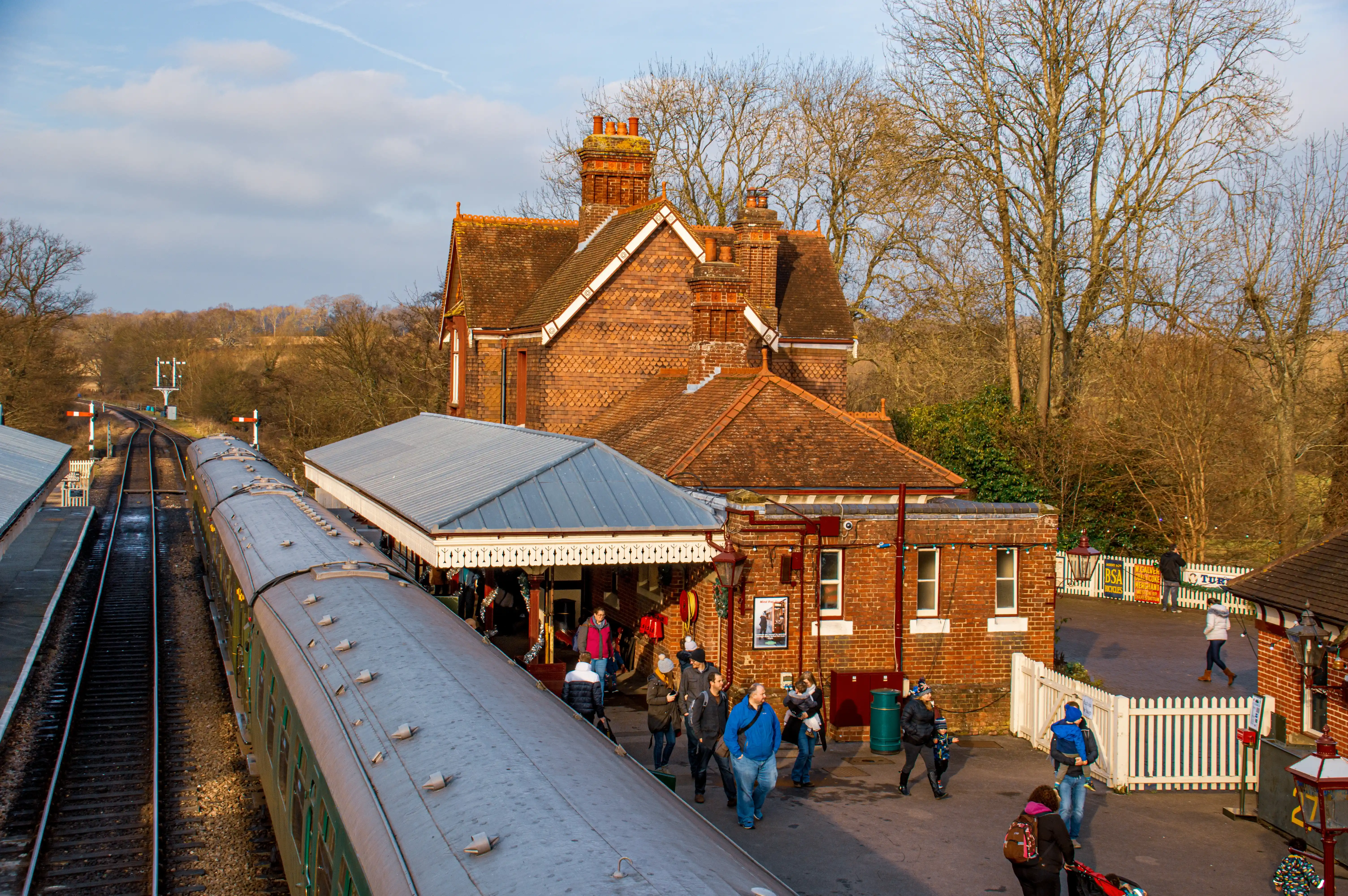 A train waits at a picturesque, old-fashioned brick railway station. People are seen on the platform, some near the train, others walking towards the station building. The station has a classic Victorian design with tiled roofs and decorative eaves. The surrounding area features trees and railway tracks.