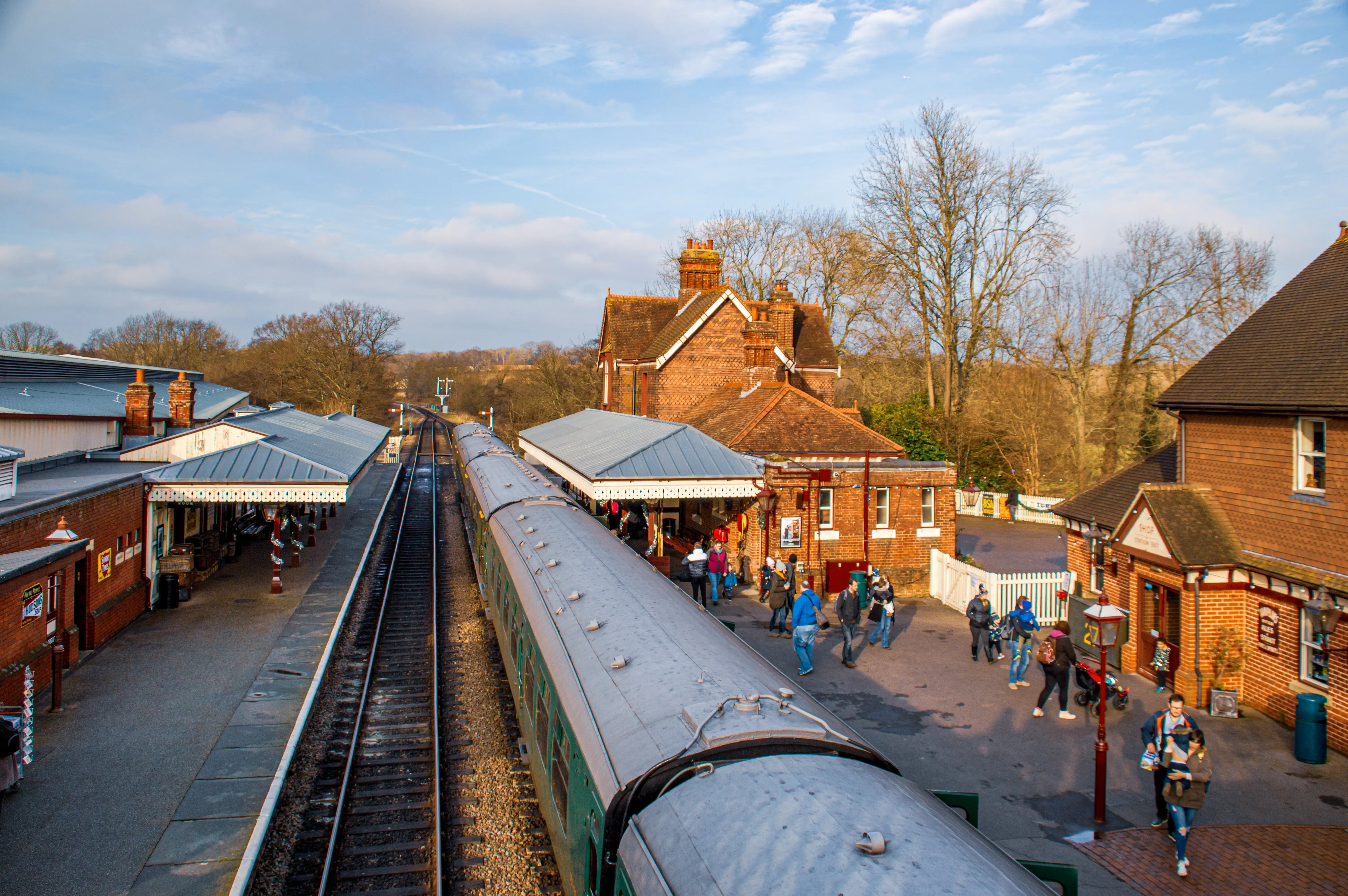 A train waits at a picturesque, old-fashioned brick railway station. People are seen on the platform, some near the train, others walking towards the station building. The station has a classic Victorian design with tiled roofs and decorative eaves. The surrounding area features trees and railway tracks.