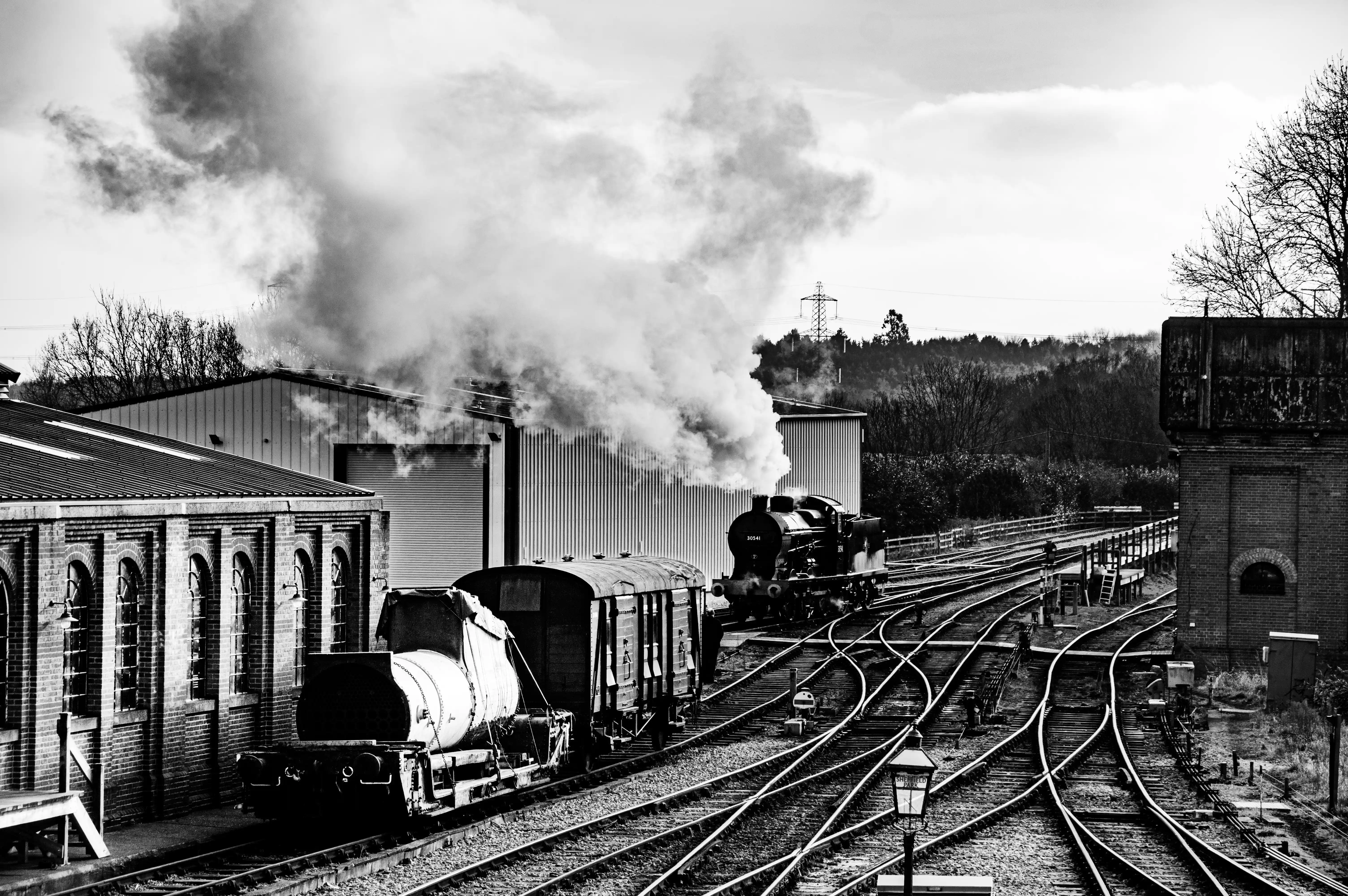 A train waits at a picturesque, old-fashioned brick railway station. People are seen on the platform, some near the train, others walking towards the station building. The station has a classic Victorian design with tiled roofs and decorative eaves. The surrounding area features trees and railway tracks.