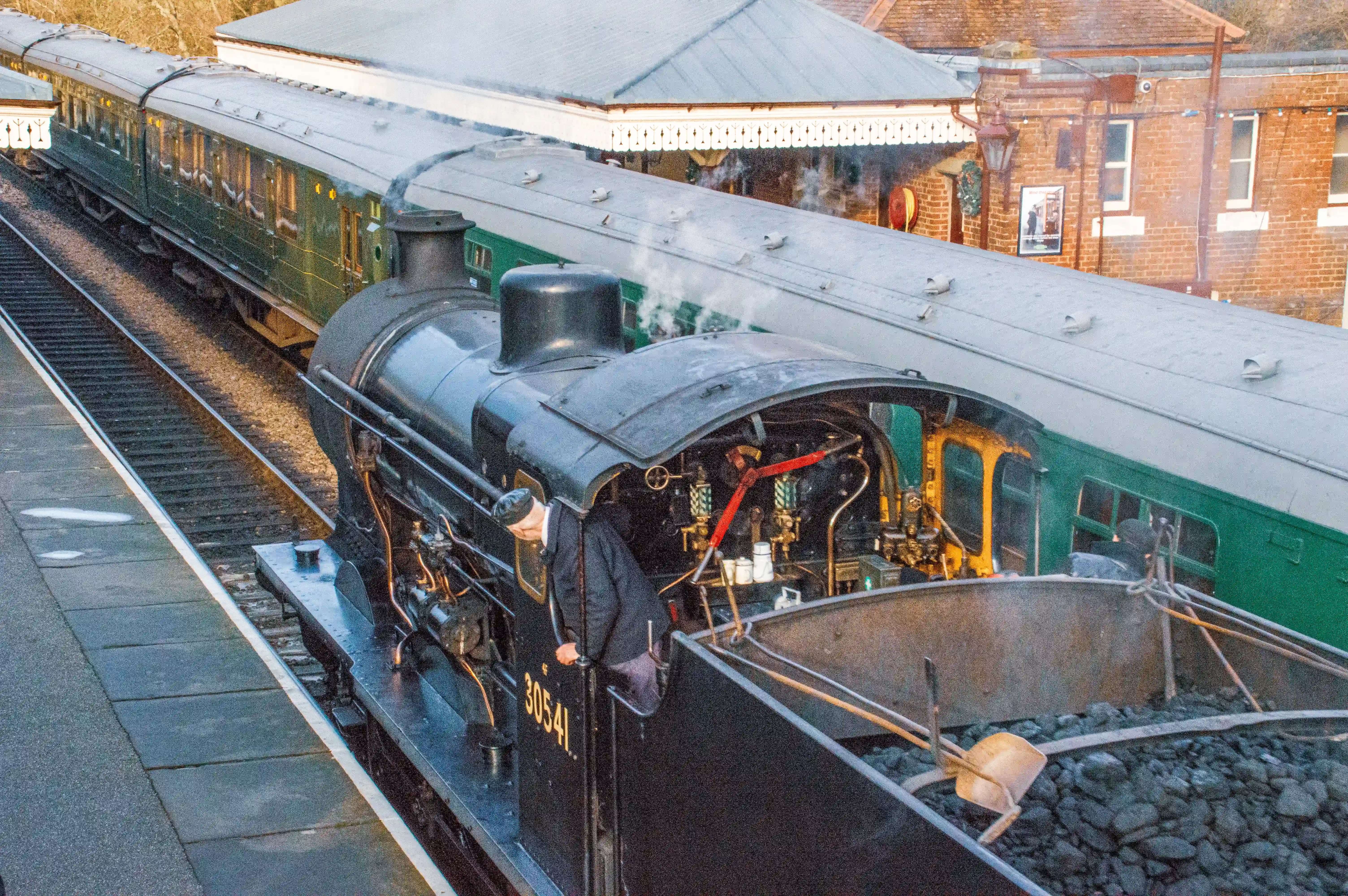 A steam locomotive numbered 30541, with a man in the cab, sits at a train station next to a green carriage, with a coal tender full of fuel.
