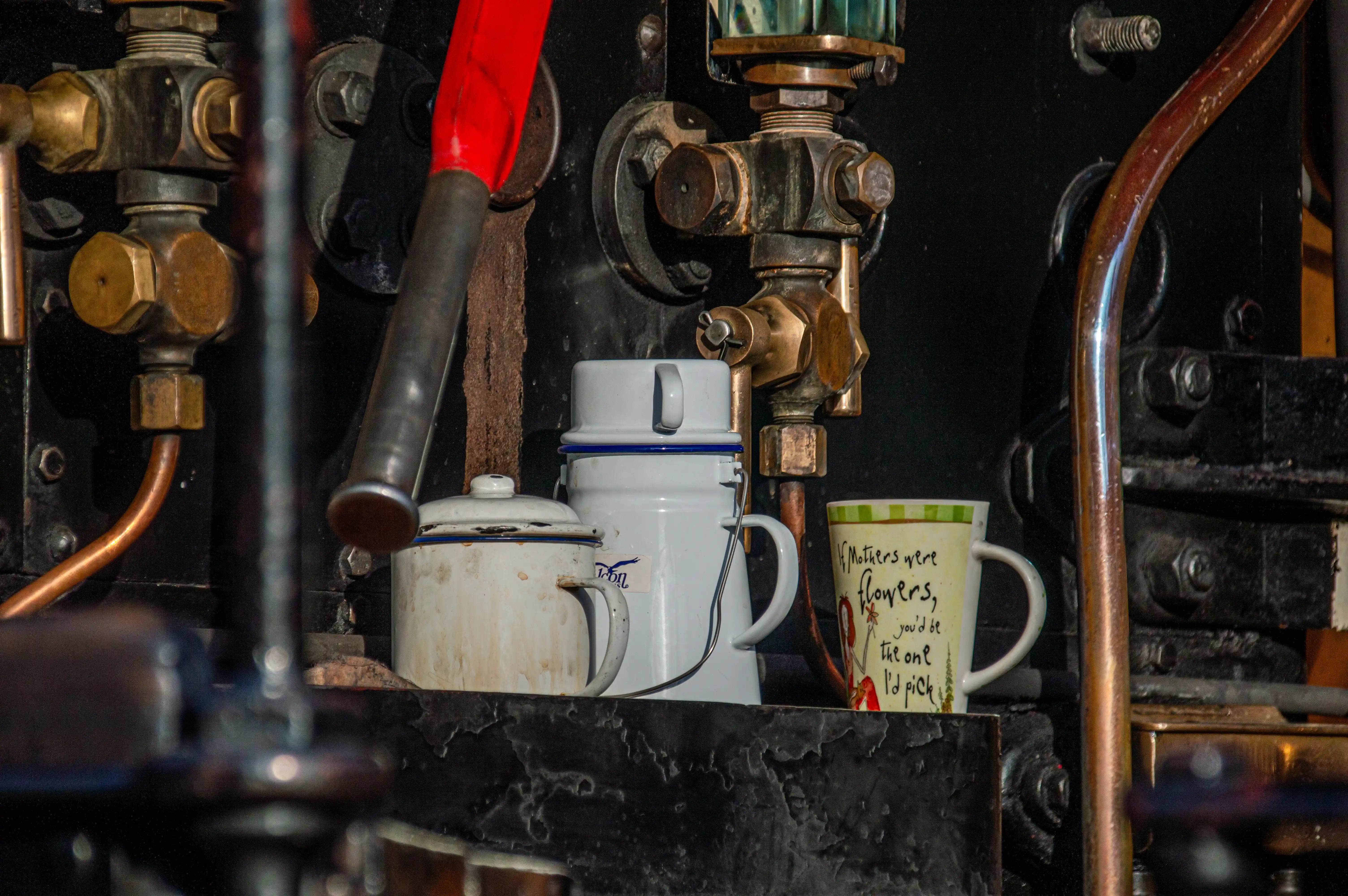 A close-up view of two white enamel jugs and a mug with a floral quote, positioned on a shelf in front of the intricate pipework and valves of a steam engine.