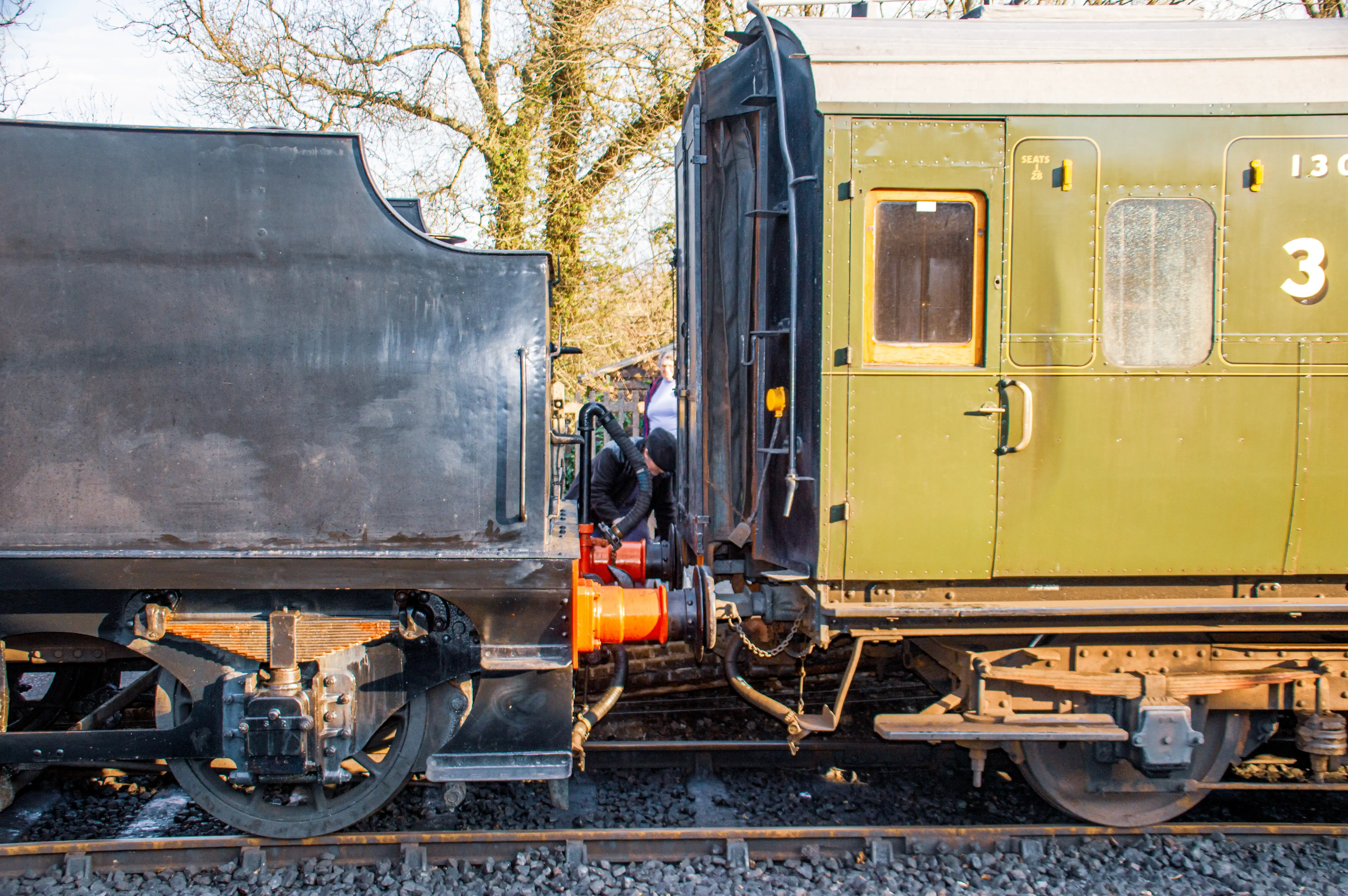 Close-up side view of a black steam locomotive tender coupled to a green heritage passenger carriage. A worker is seen between them, attending to the coupling mechanism.
