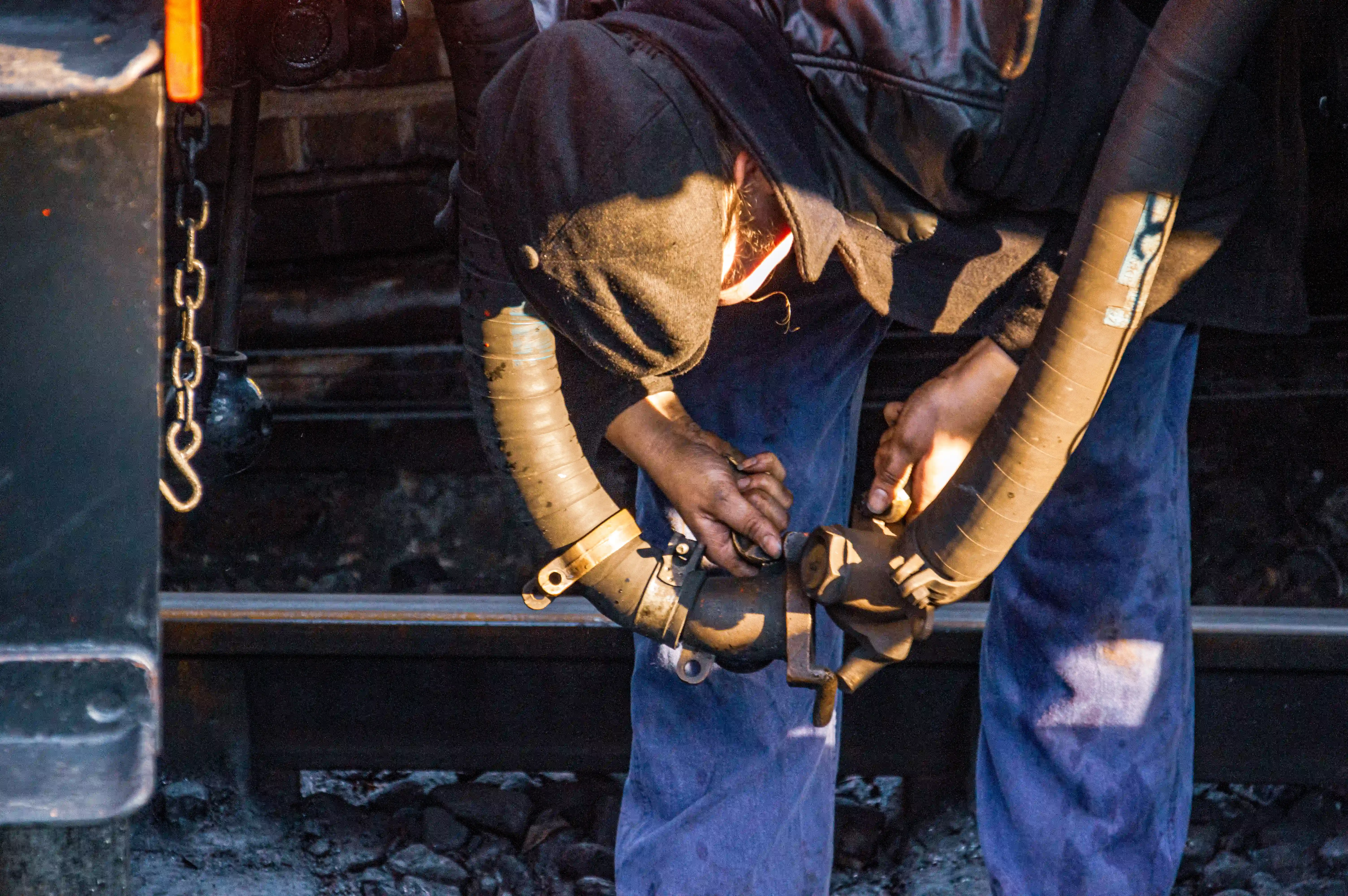 A person wearing a black hooded top and blue overalls connects a thick, ribbed hose with metallic fittings. Their hands are visible, engaged in the connection. In the background, there are train tracks, metal structures, and loose rubble.