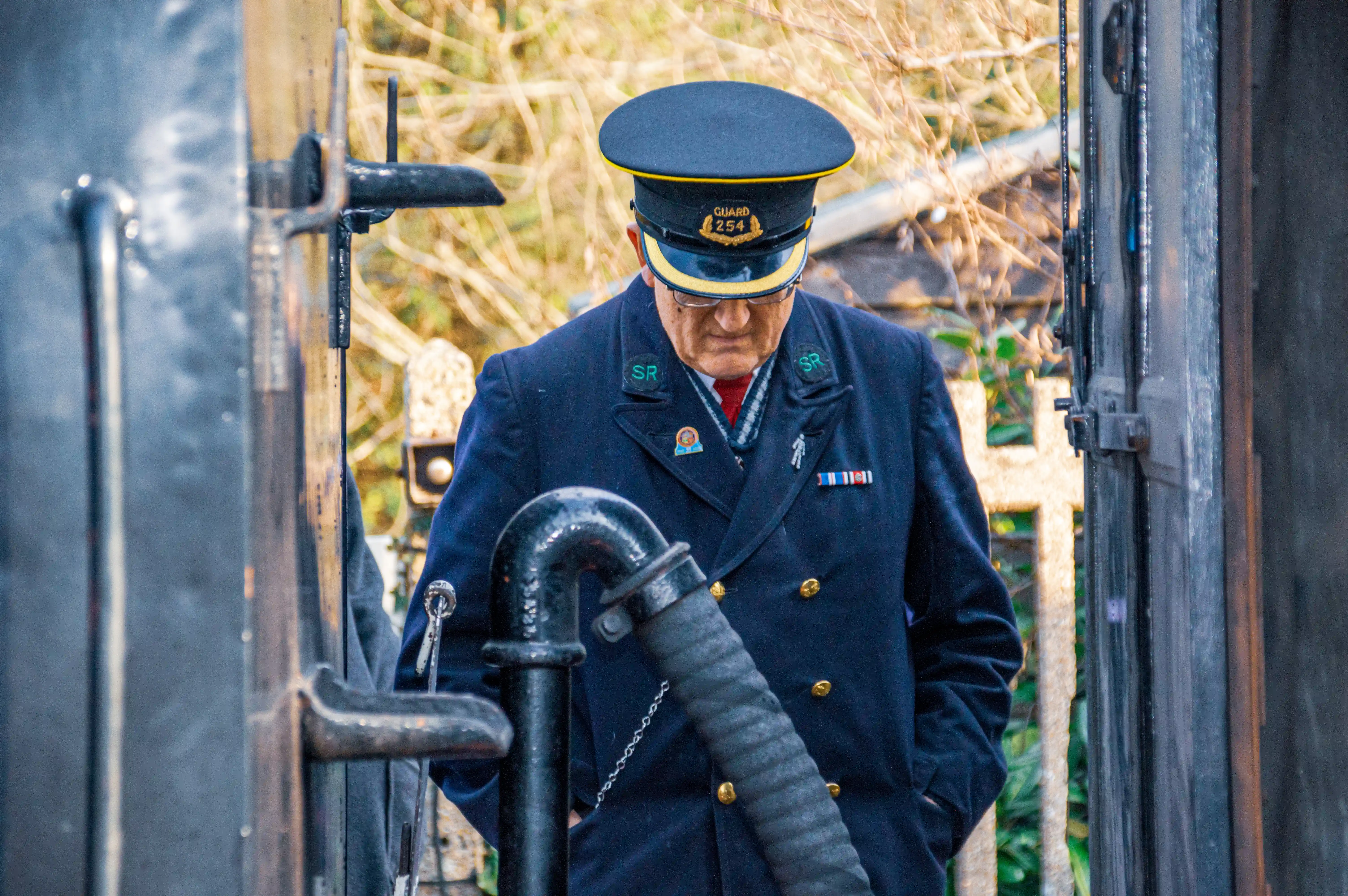 A train guard in a dark blue uniform and peaked cap looks down. The cap has a badge that reads GUARD 254. The uniform has SR embroidered on the collars and several medals or ribbons on the chest. He is standing next to a steam engine with metal pipes and railings visible.
