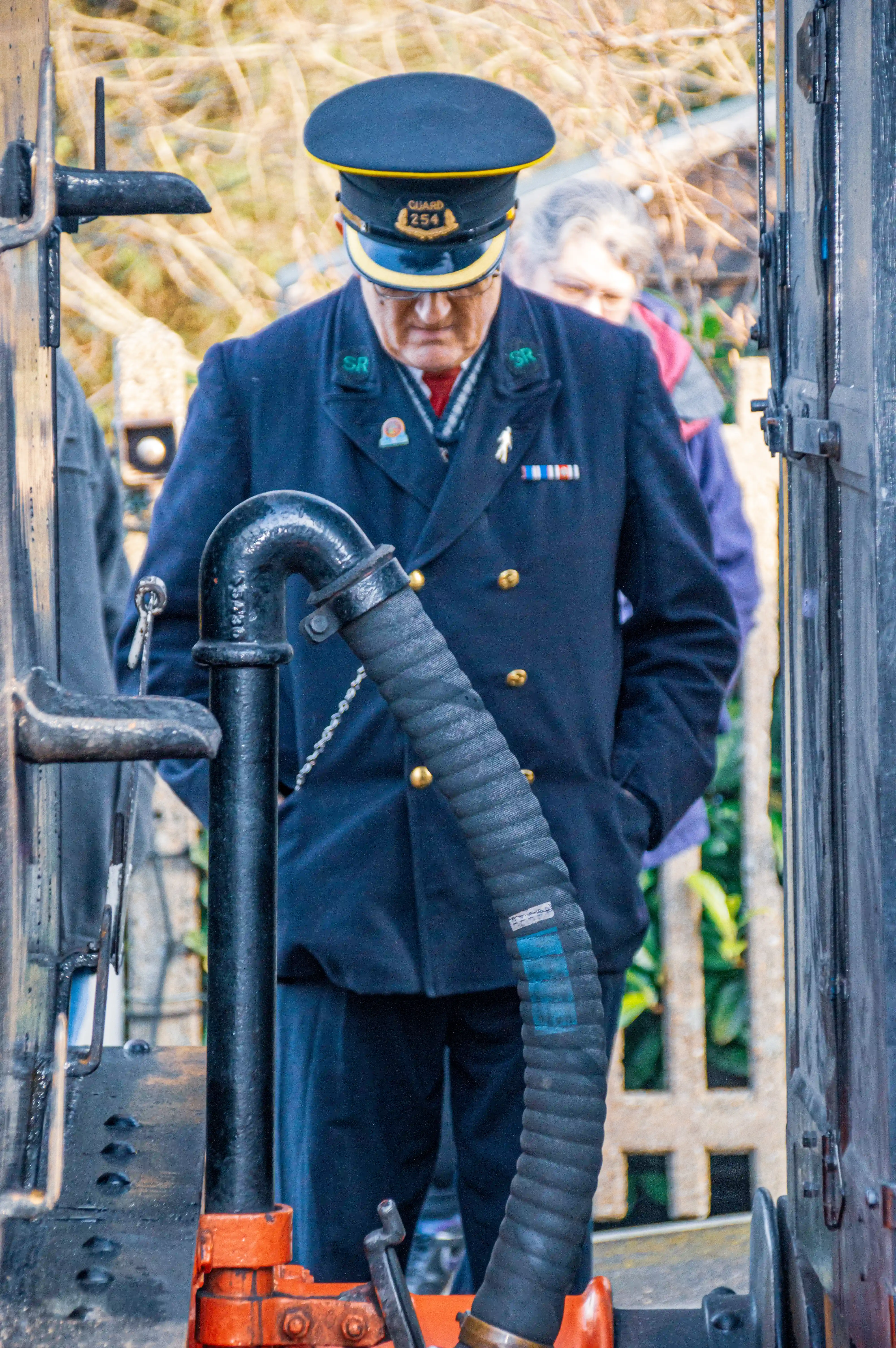 A train guard in a dark blue uniform and peaked cap looks down. The cap has a badge that reads 