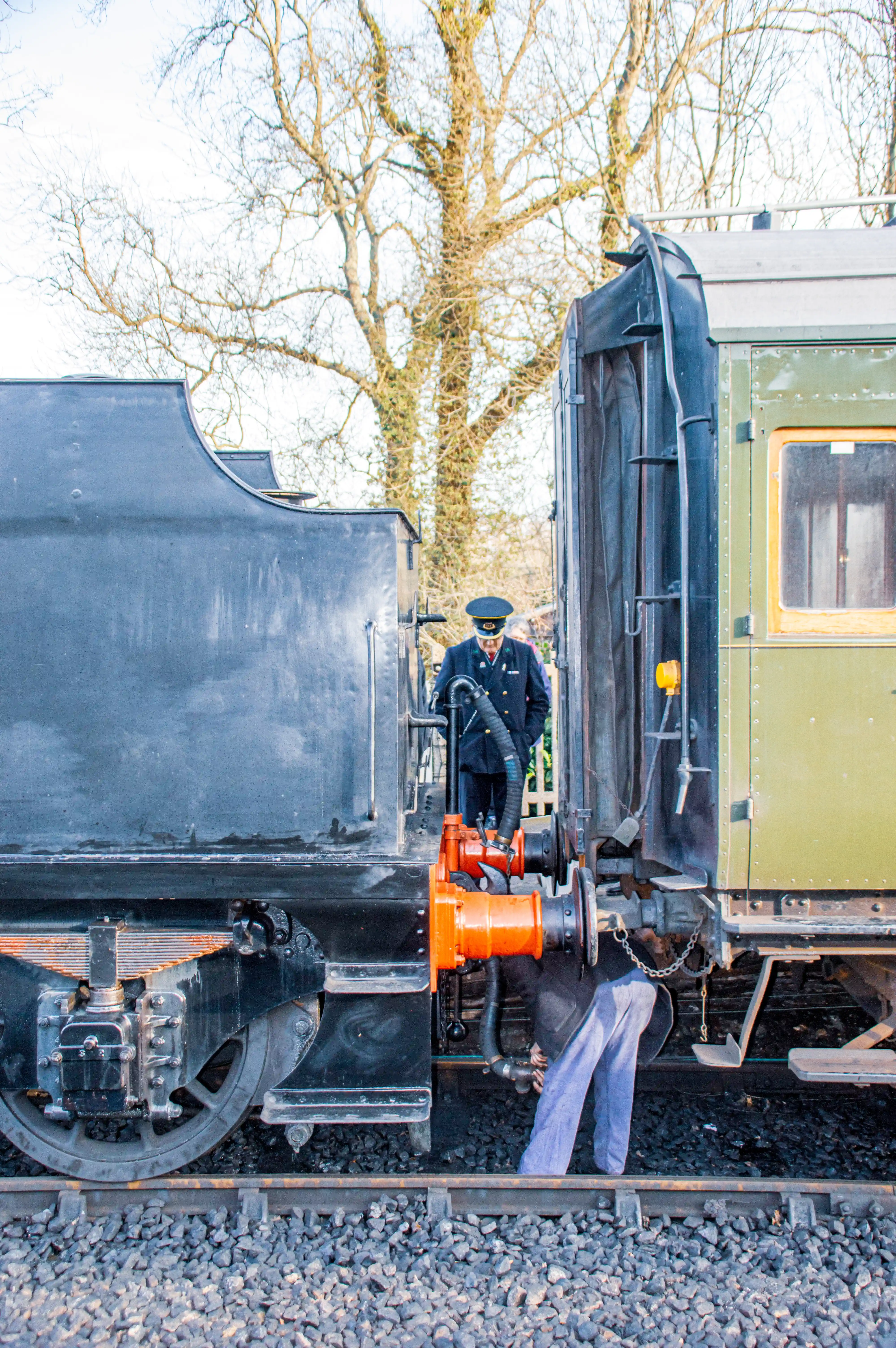 Close-up side view of a black steam locomotive tender coupled to a green heritage passenger carriage. A worker is seen between them, attending to the coupling mechanism.