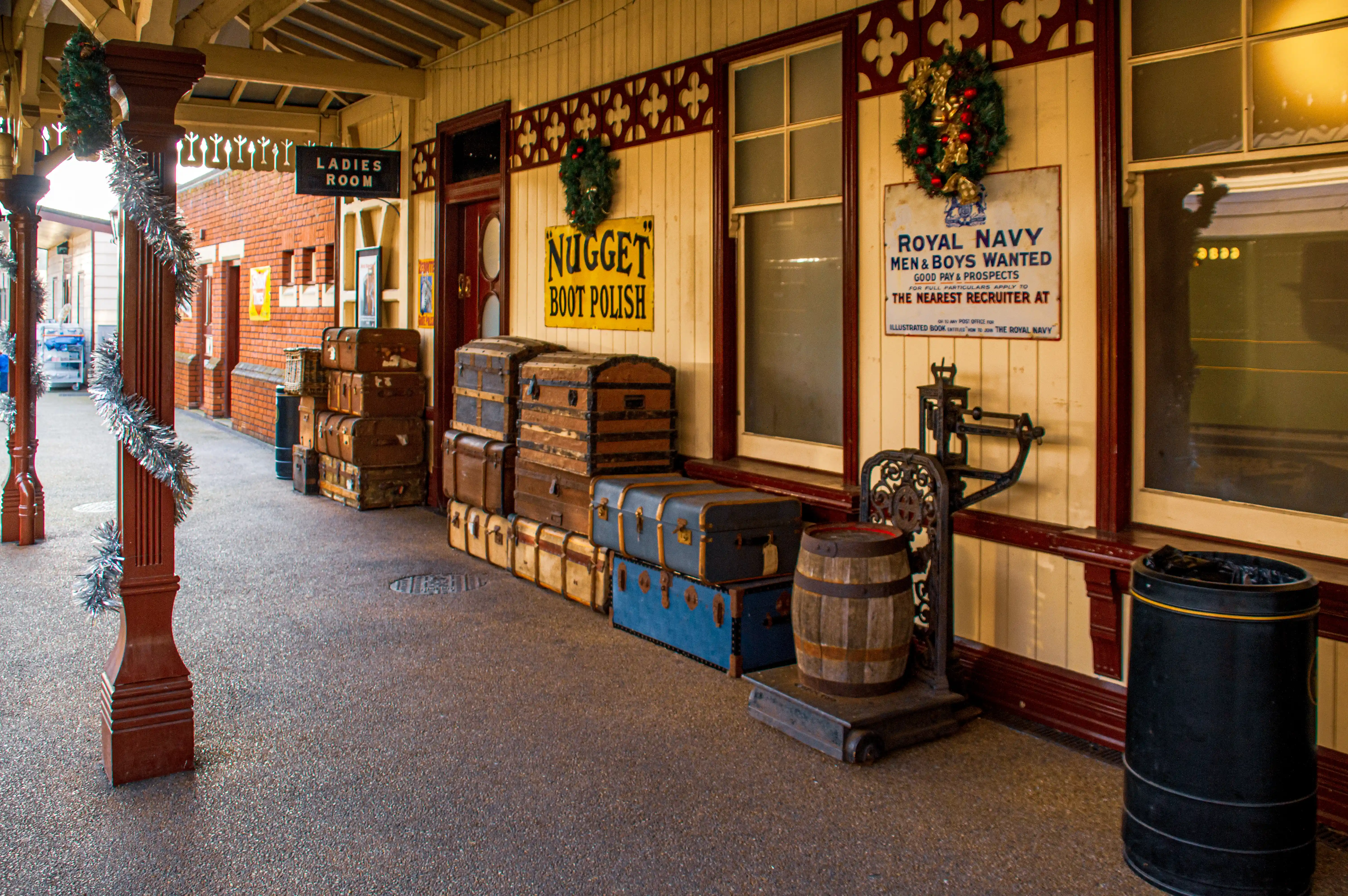 A historic train station platform decorated for Christmas, with piles of vintage suitcases, a 'Ladies Room' sign, and a wooden barrel on a weighing scale.