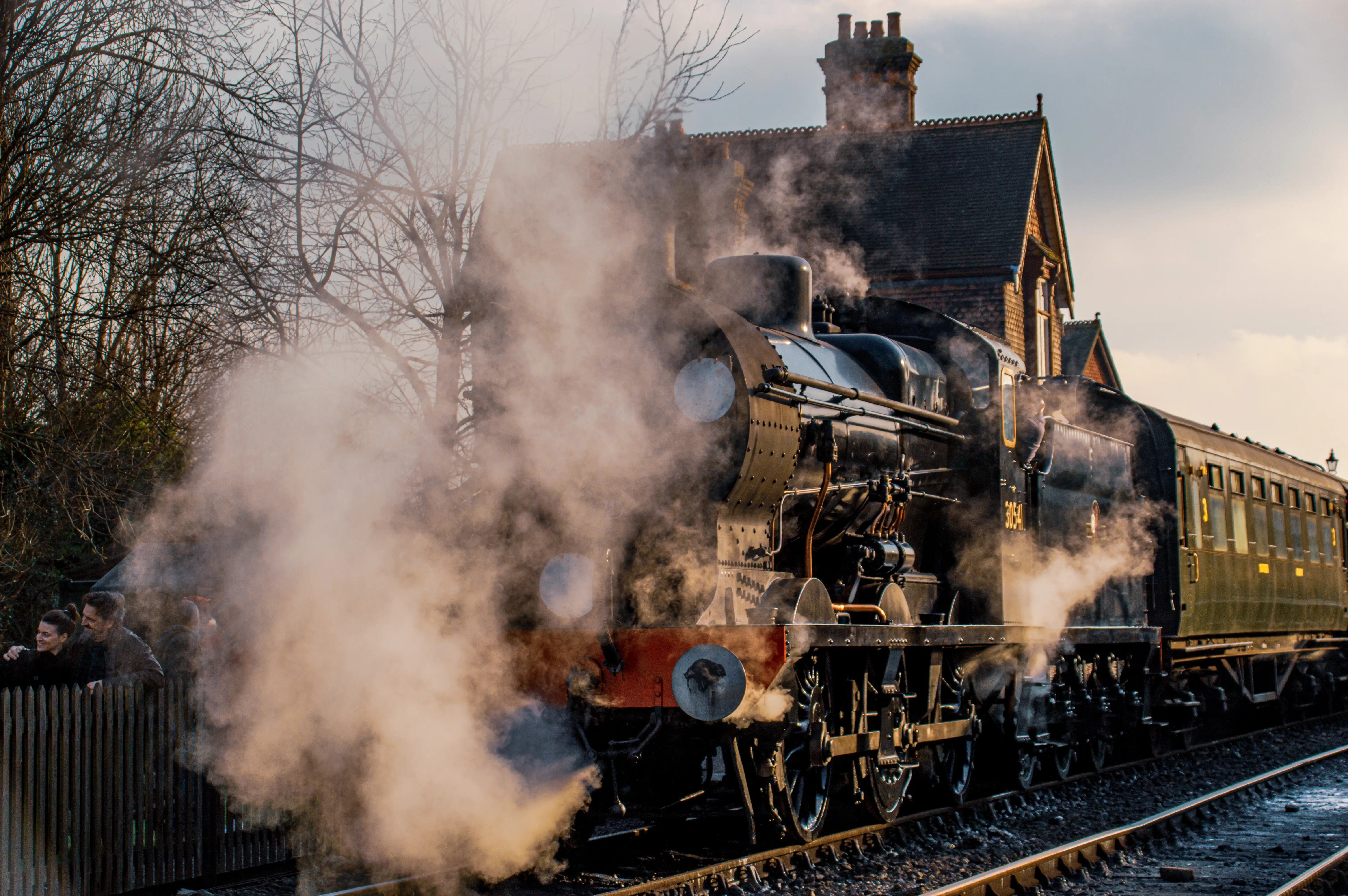 A black steam train with carriages, emitting a large cloud of white steam, is beside a traditional brick station building. People are visible behind a fence on the left.
