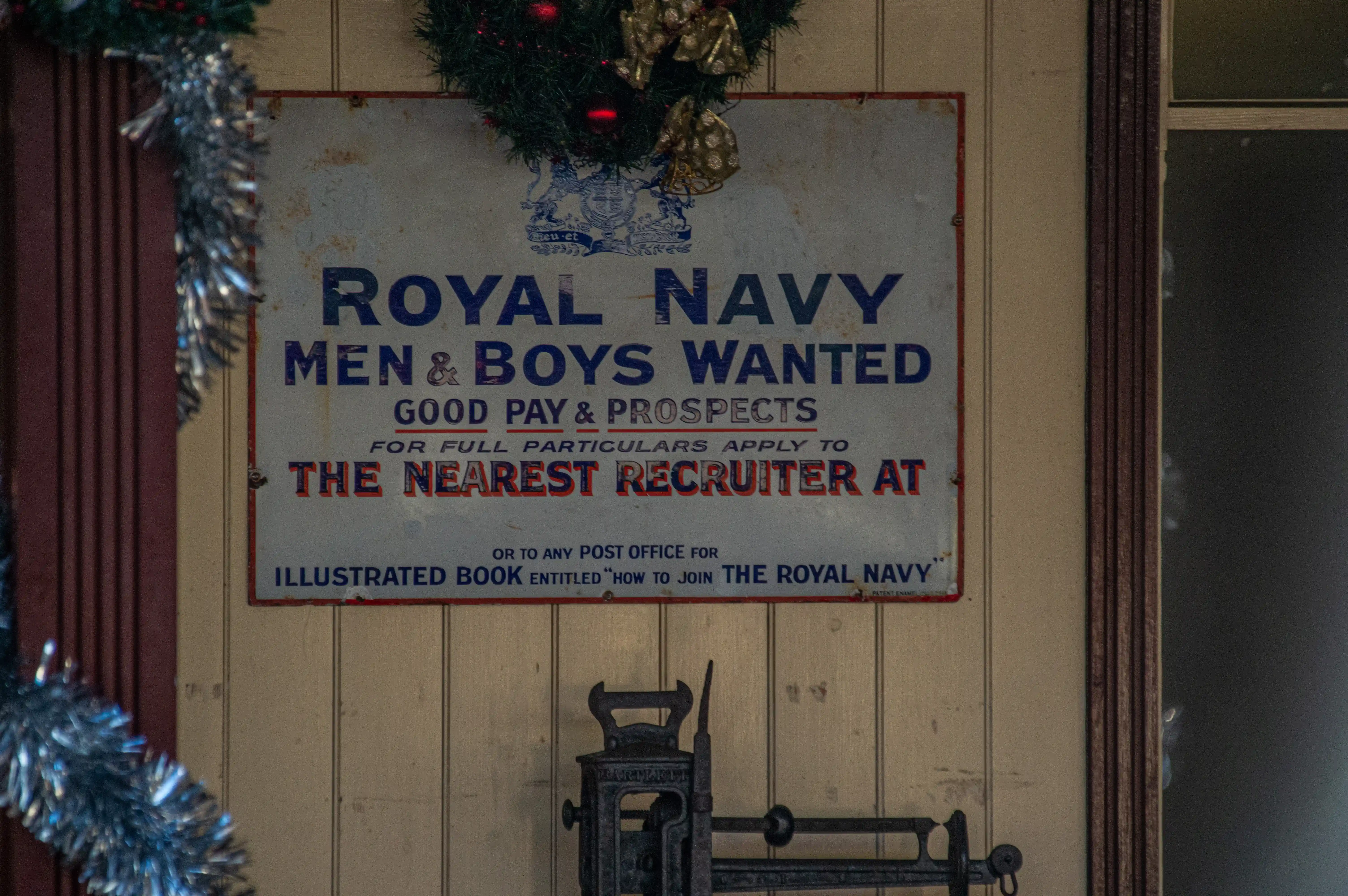 A close-up, eye-level view of a vintage Royal Navy recruitment poster. The poster is mounted on a light-coloured wooden wall and is adorned with a Christmas wreath and silver tinsel on its left side. The poster text is predominantly in dark blue and red, with the headline ROYAL NAVY MEN & BOYS WANTED clearly visible. Below this, it states GOOD PAY & PROSPECTS and instructs potential recruits to APPLY TO THE NEAREST RECRUITER AT. A lower section mentions obtaining an ILLUSTRATED BOOK ENTITLED 'HOW TO JOIN THE ROYAL NAVY' from any post office. A vintage scale mechanism is visible at the bottom of the frame.
