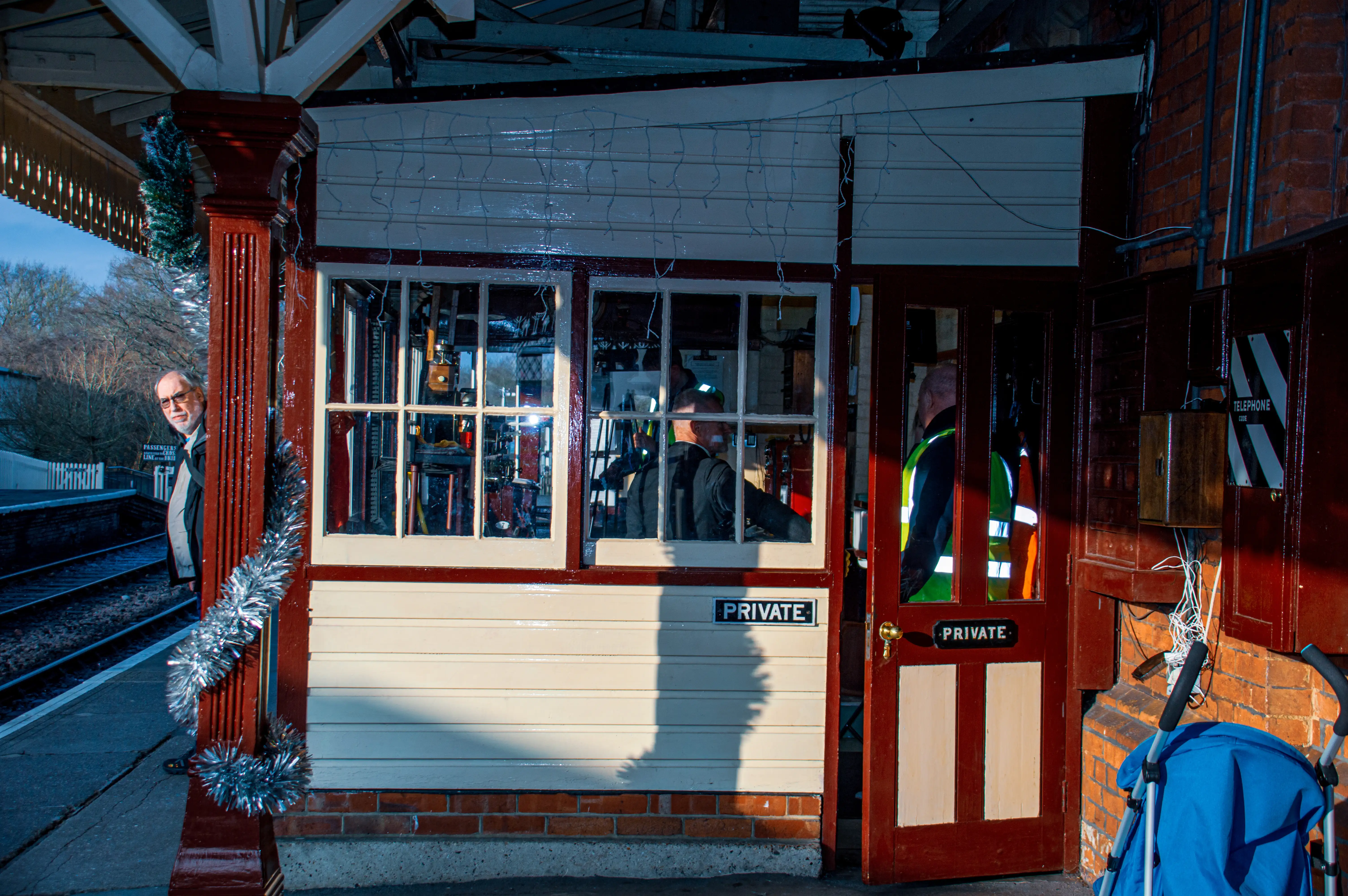 A man with glasses and a grey jacket peeks from behind a red column decorated with silver tinsel at a quaint train station. The station booth has large windows showing people inside, and a PRIVATE sign is on the wall. A second PRIVATE sign is on a door, and a blue pushchair is visible to the right.