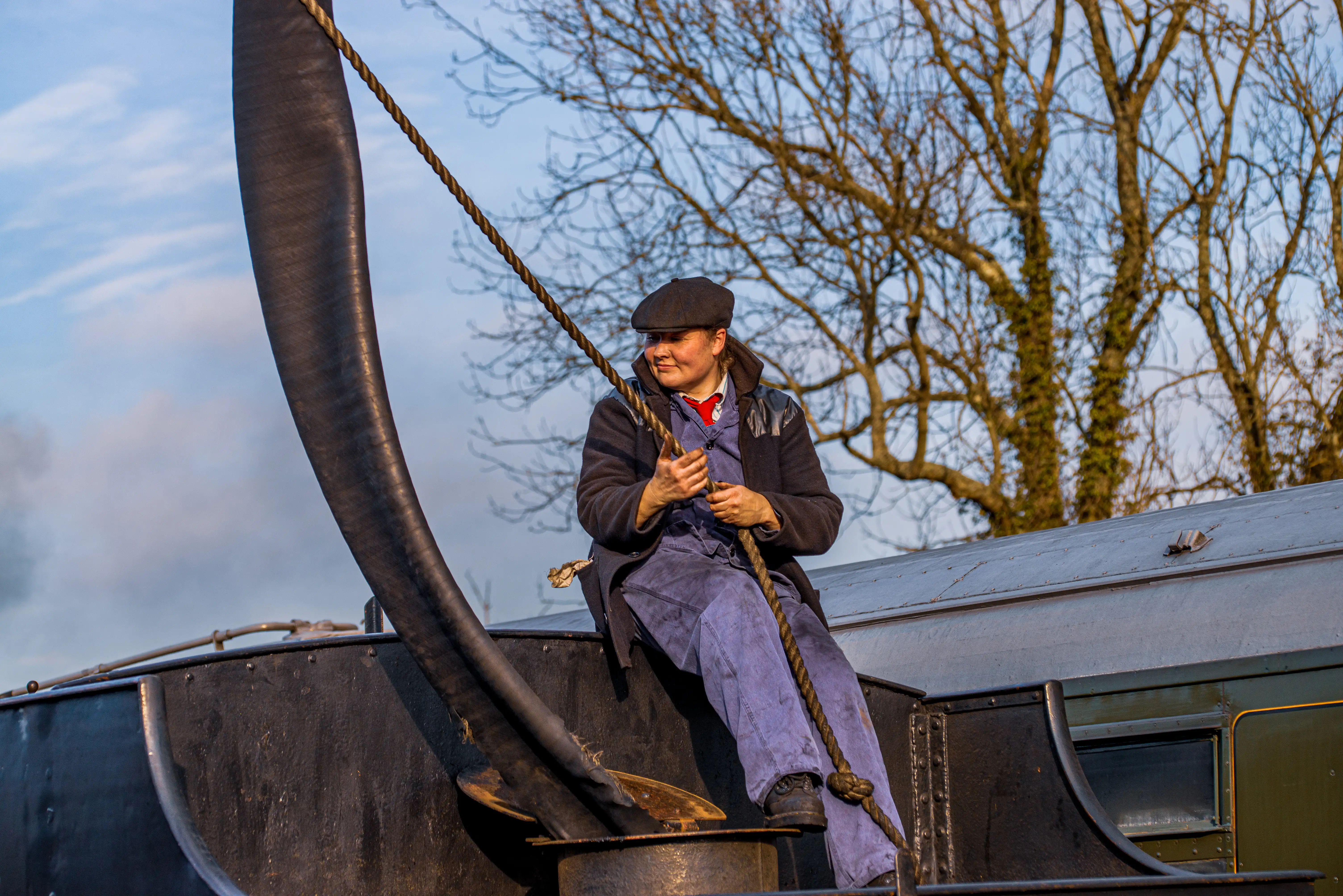 Railway worker sitting on a steam locomotive holding a rope attached to a large hose on a cool day with bare trees behind.