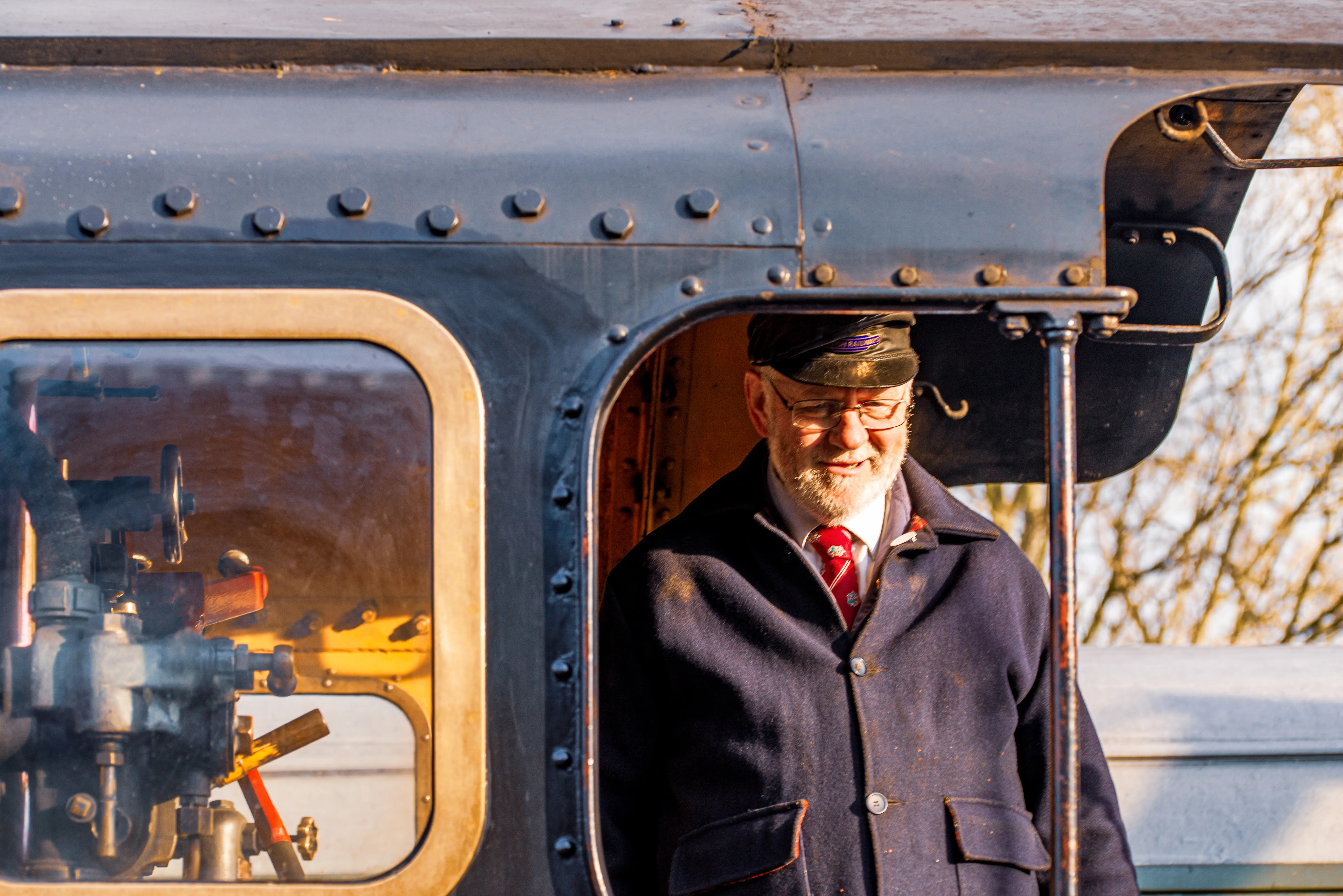 A middle-aged man with a white beard and glasses, wearing a dark blue coat and a cap with a badge, smiles warmly. He is standing inside the cab of a steam train, with the engine's controls visible through a window on the left.