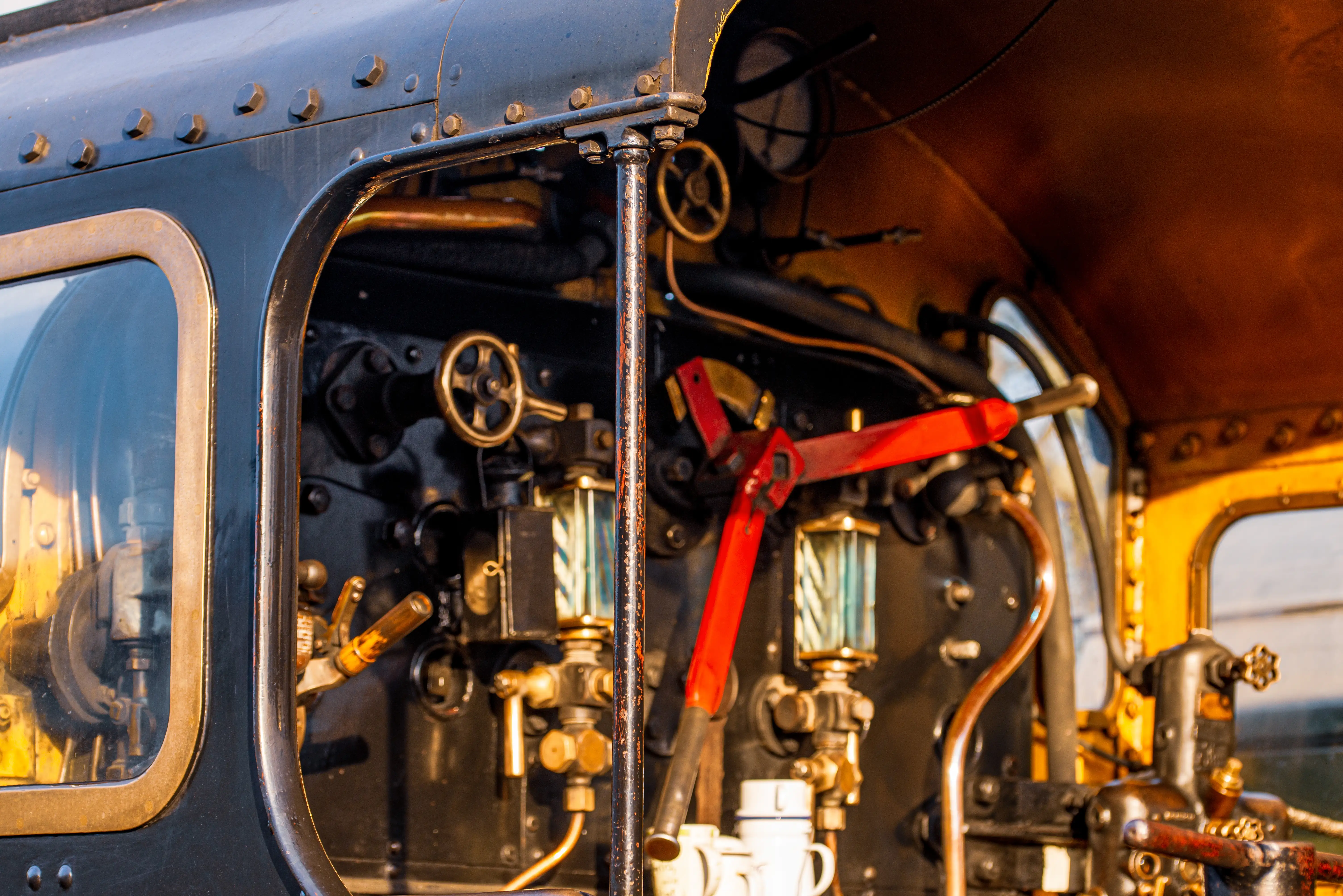 Interior view of a steam train's control panel, featuring brass levers, gauges with liquid, and copper piping.
