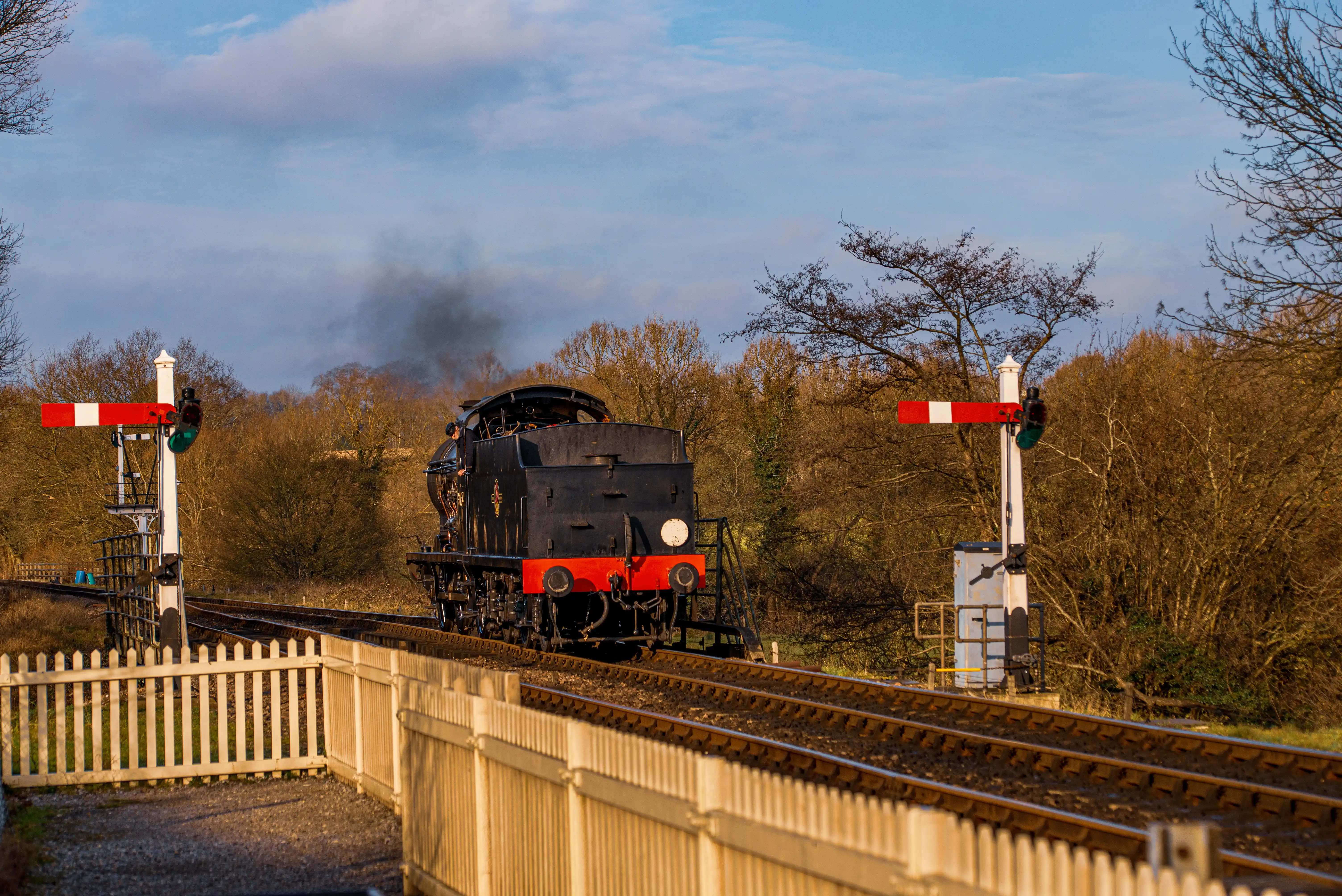 A black steam train with red accents on the front travels on a track, emitting black smoke. Two railway signals with red and white arms are visible. A white wooden fence is in the foreground.