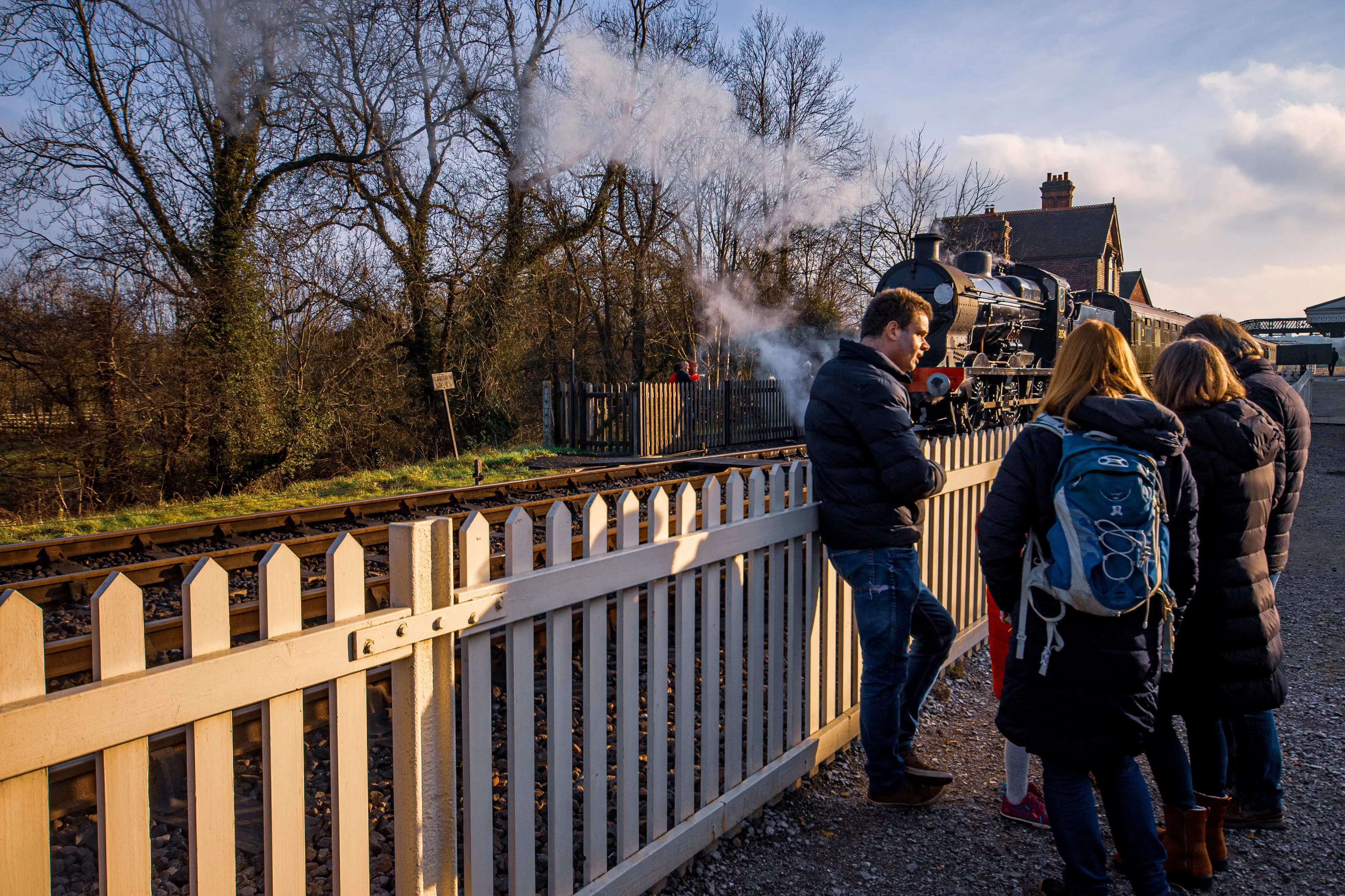 Several people are gathered at a railway station, looking towards a vintage steam train billowing smoke. They stand behind a white wooden fence as the train pulls into the platform.