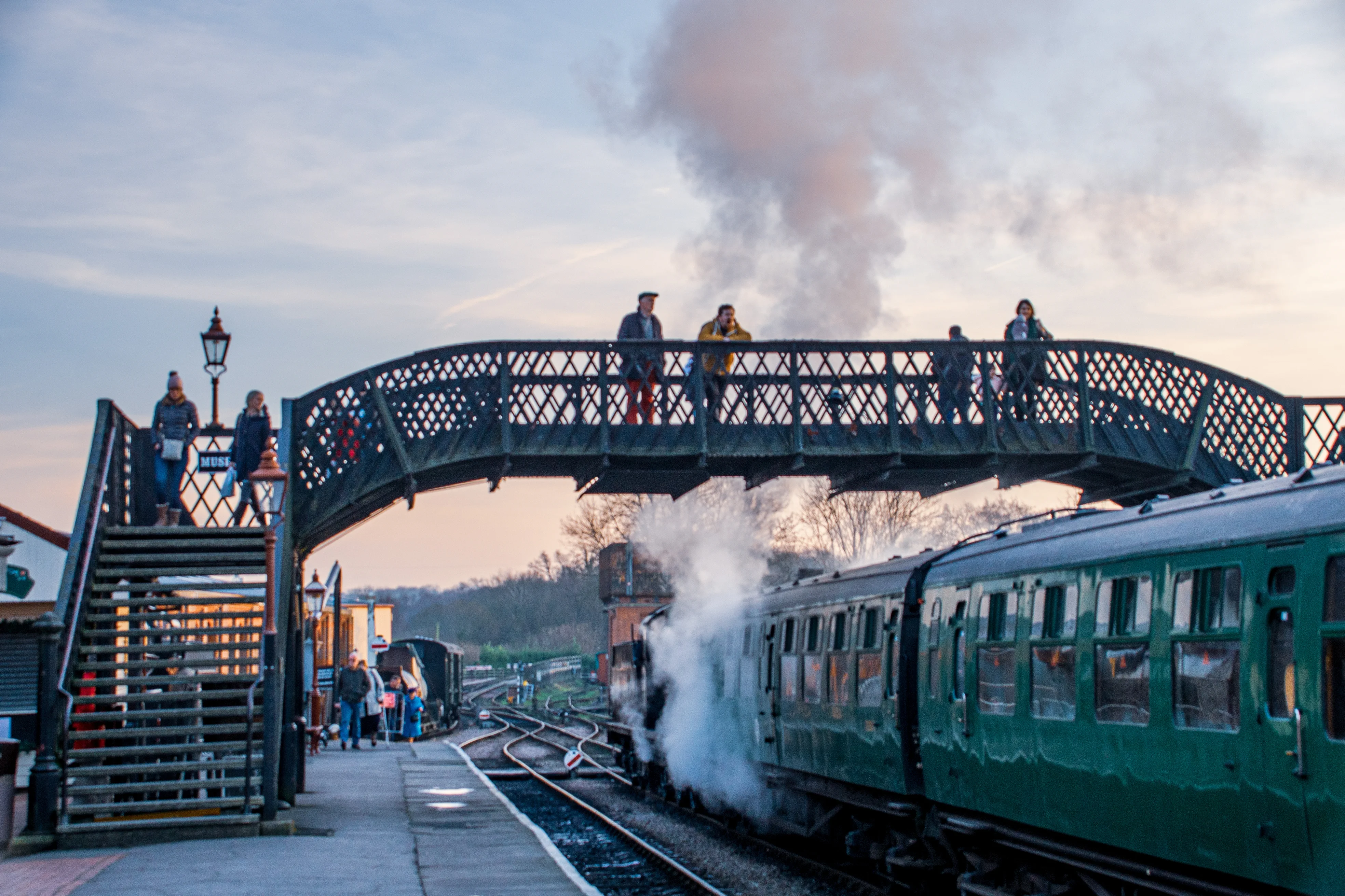 A steam train pulling into a station, with people on a platform and standing on a decorative iron footbridge overhead.