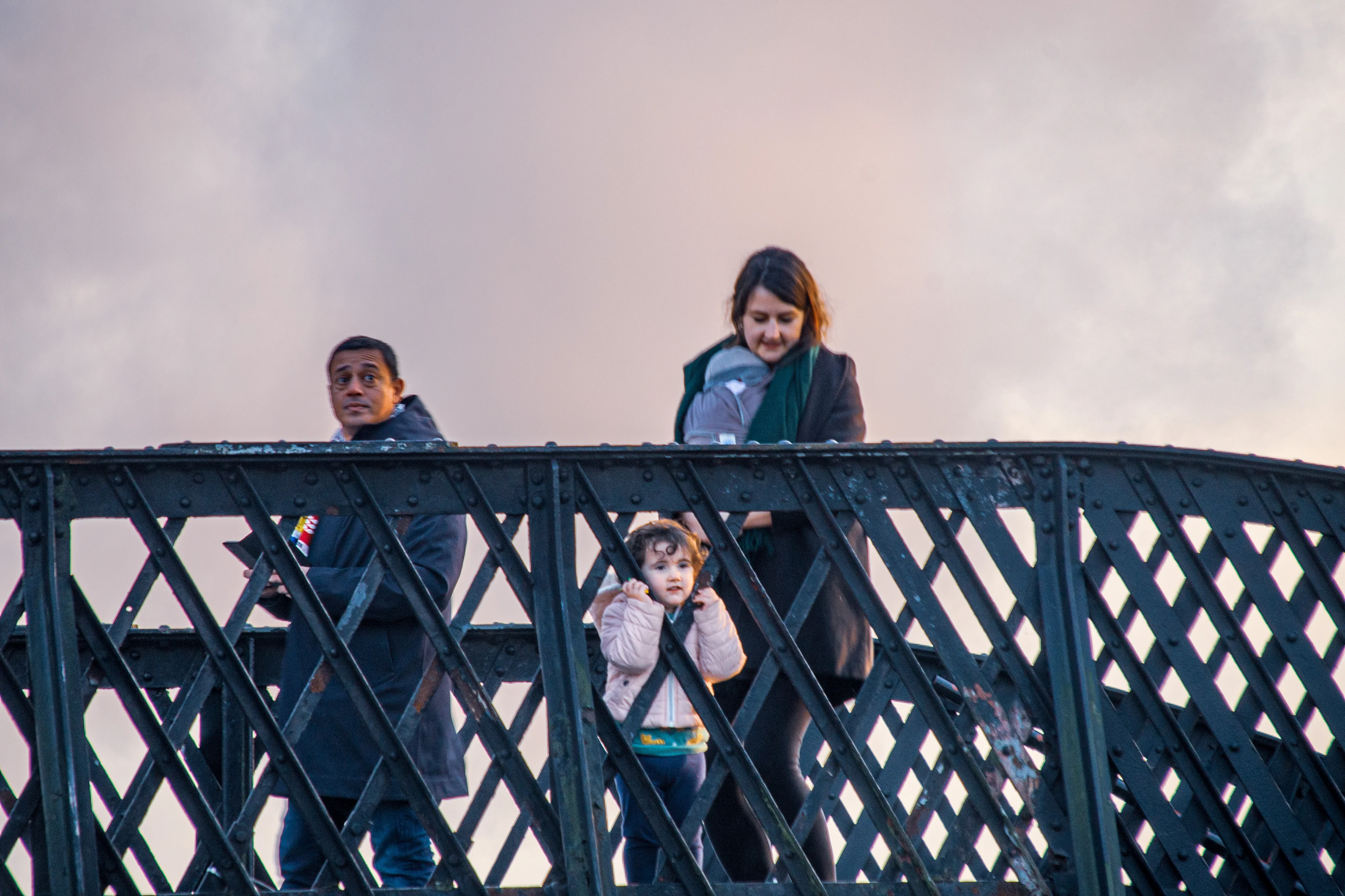 A man, a woman holding a baby in a carrier, and a small child stand on a metal bridge railing against a cloudy sky.