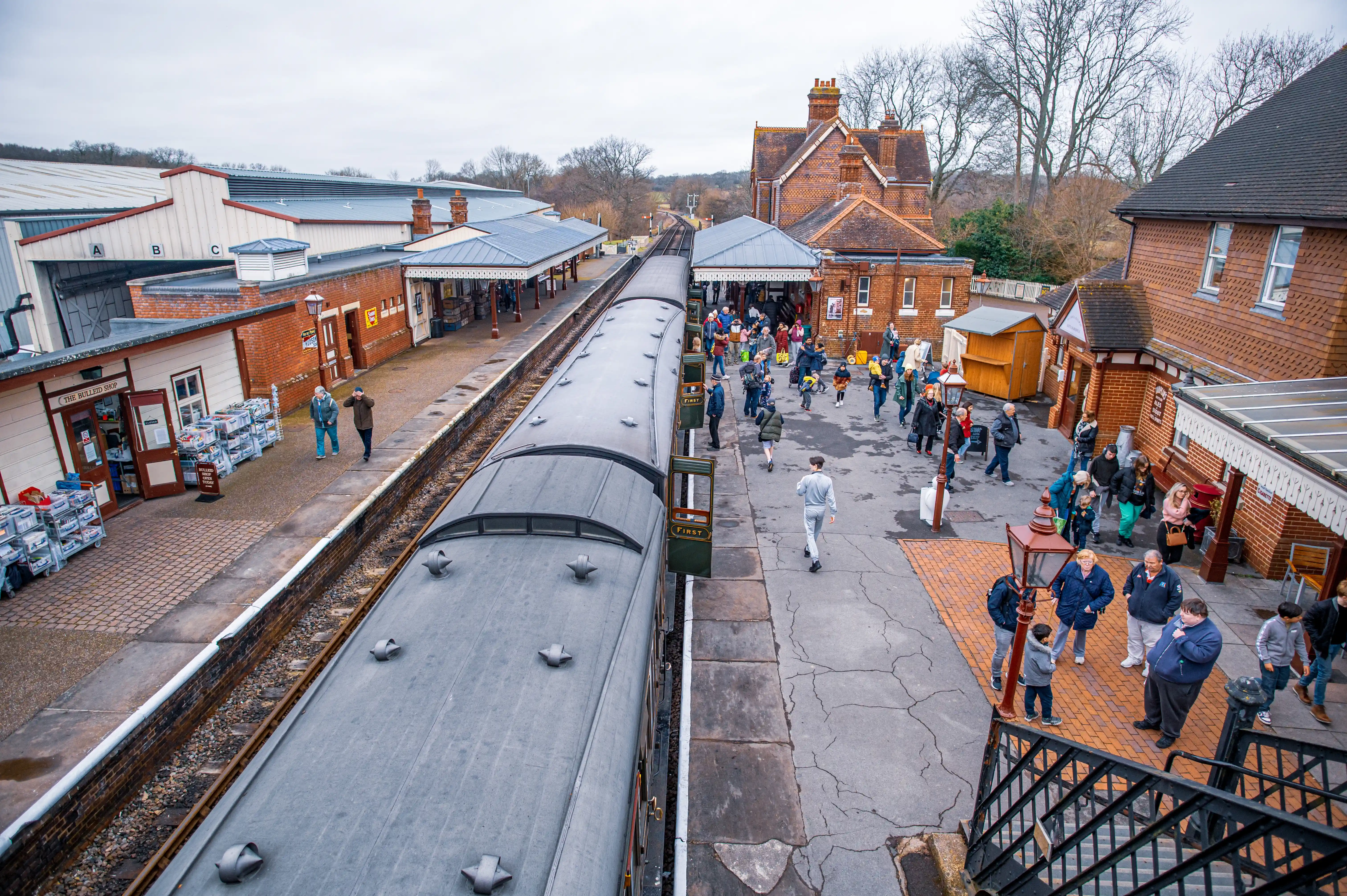 An elevated view of a vintage train at a platform in a traditional brick railway station, with many people standing on the platform, browsing shops, and waiting for the train under a cloudy sky.