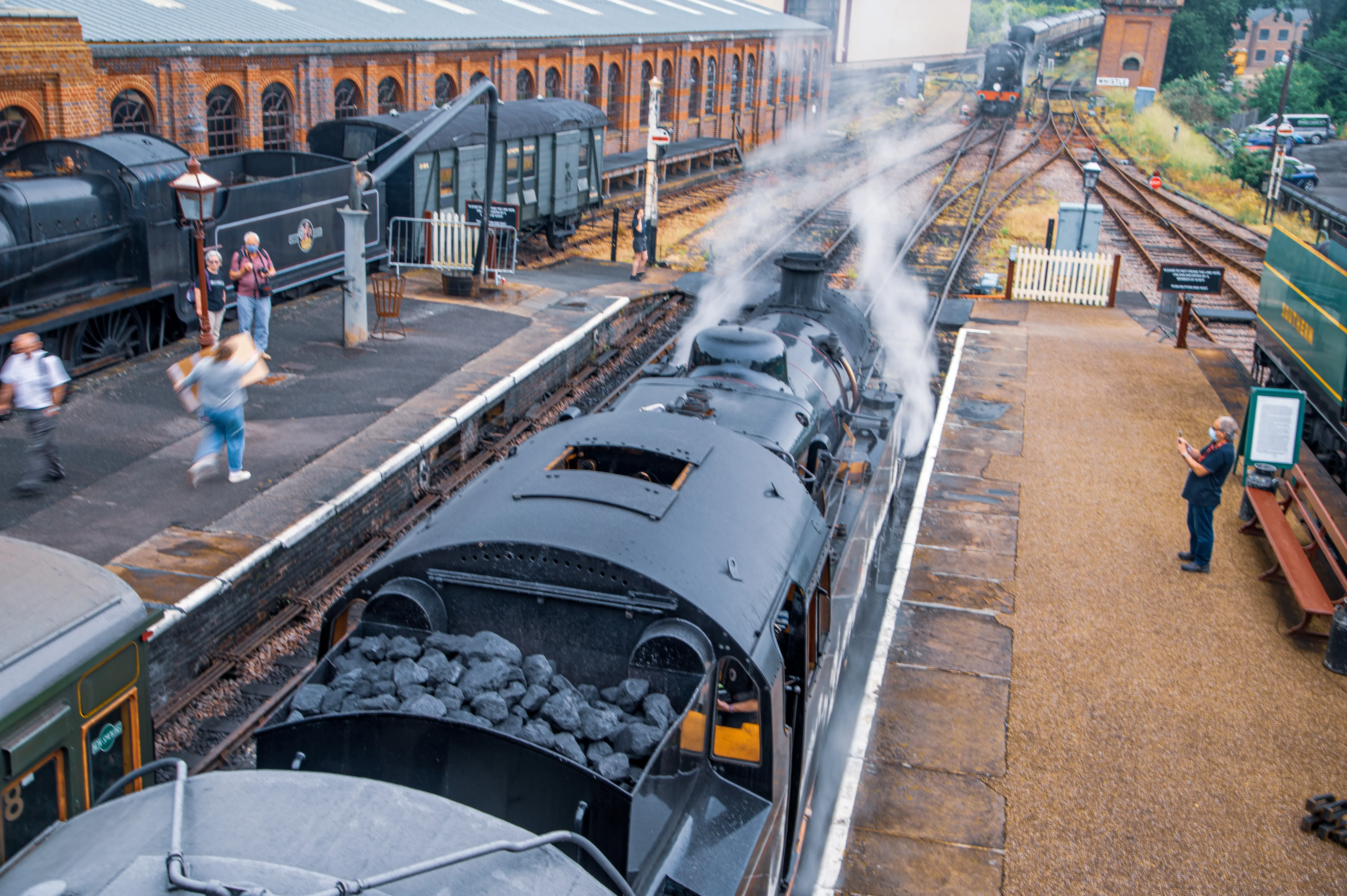 An overhead view of a steam train station with multiple steam locomotives, train carriages, tracks, and several people on the platform, one of whom is taking a photograph of a train.