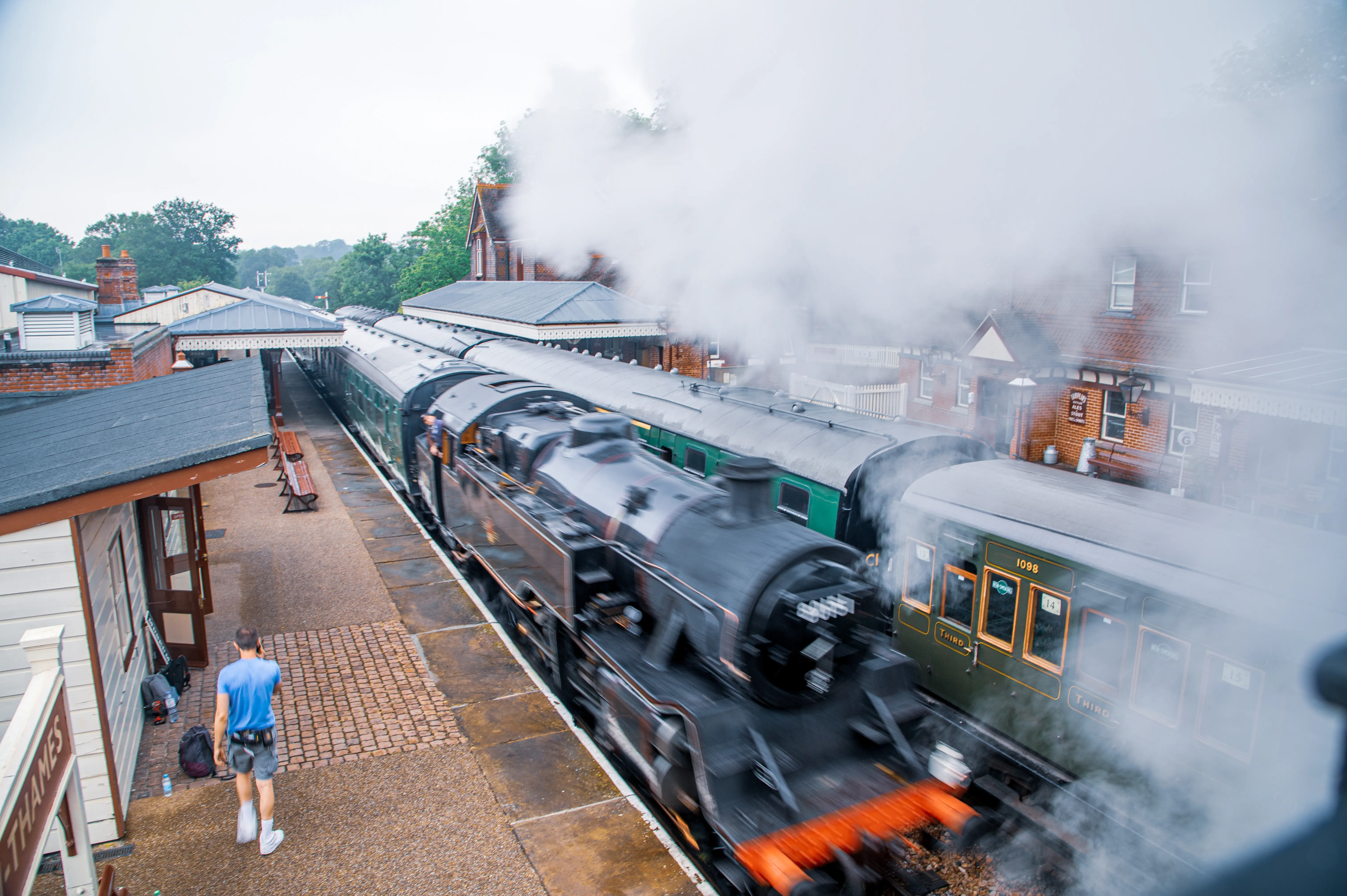 A black steam train, Number 60158, is pulling away from a station platform, billowing a large cloud of steam into the air. The train has green carriages with orange-tinted windows. A man in a blue t-shirt and grey shorts walks away from the camera on the platform.