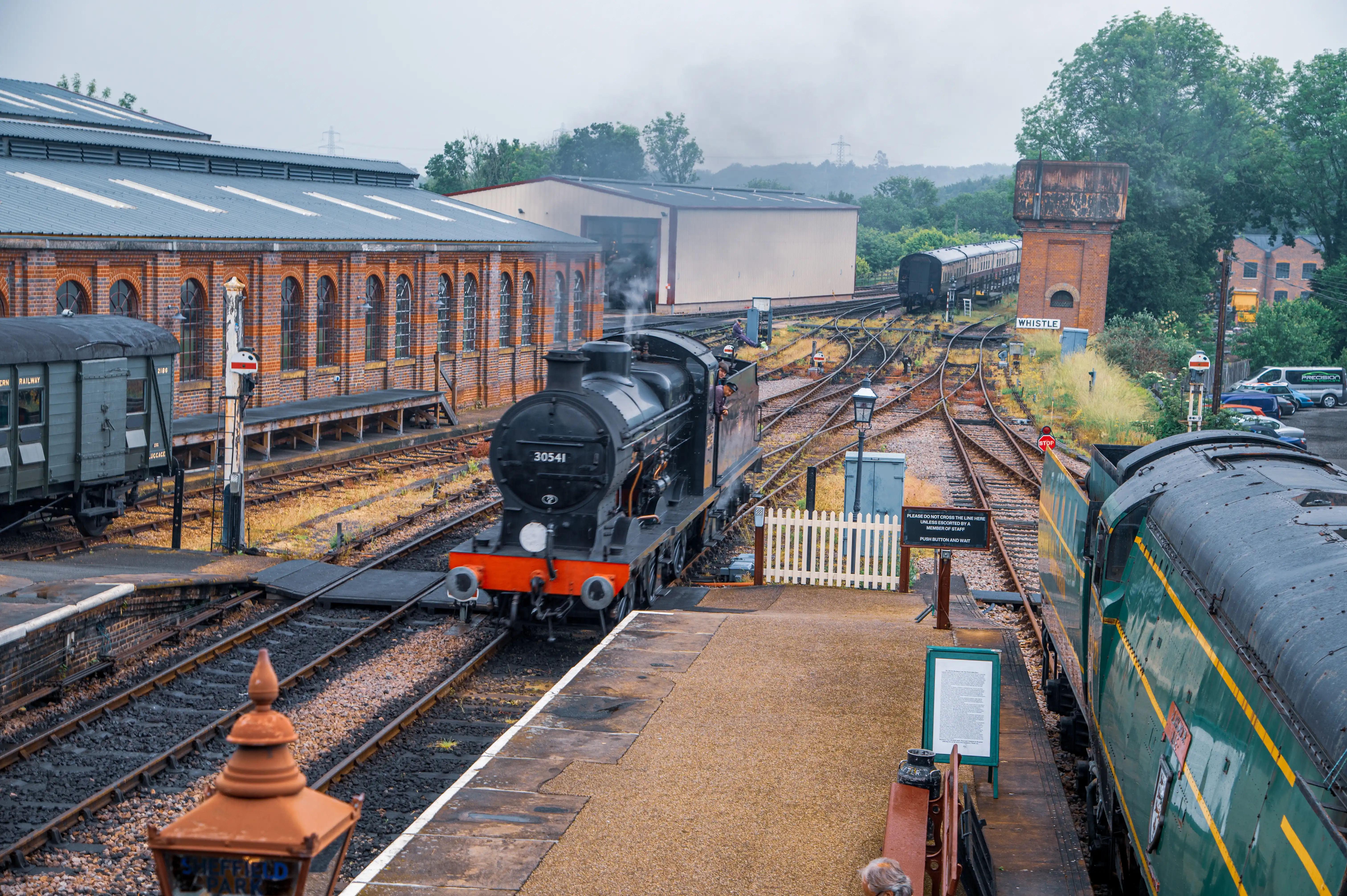 A black steam train numbered 30541 approaches a station platform, with smoke rising. A green and yellow passenger train is parked on an adjacent track. A brick building, a water tower, and various railway tracks are visible in the background under a cloudy sky.