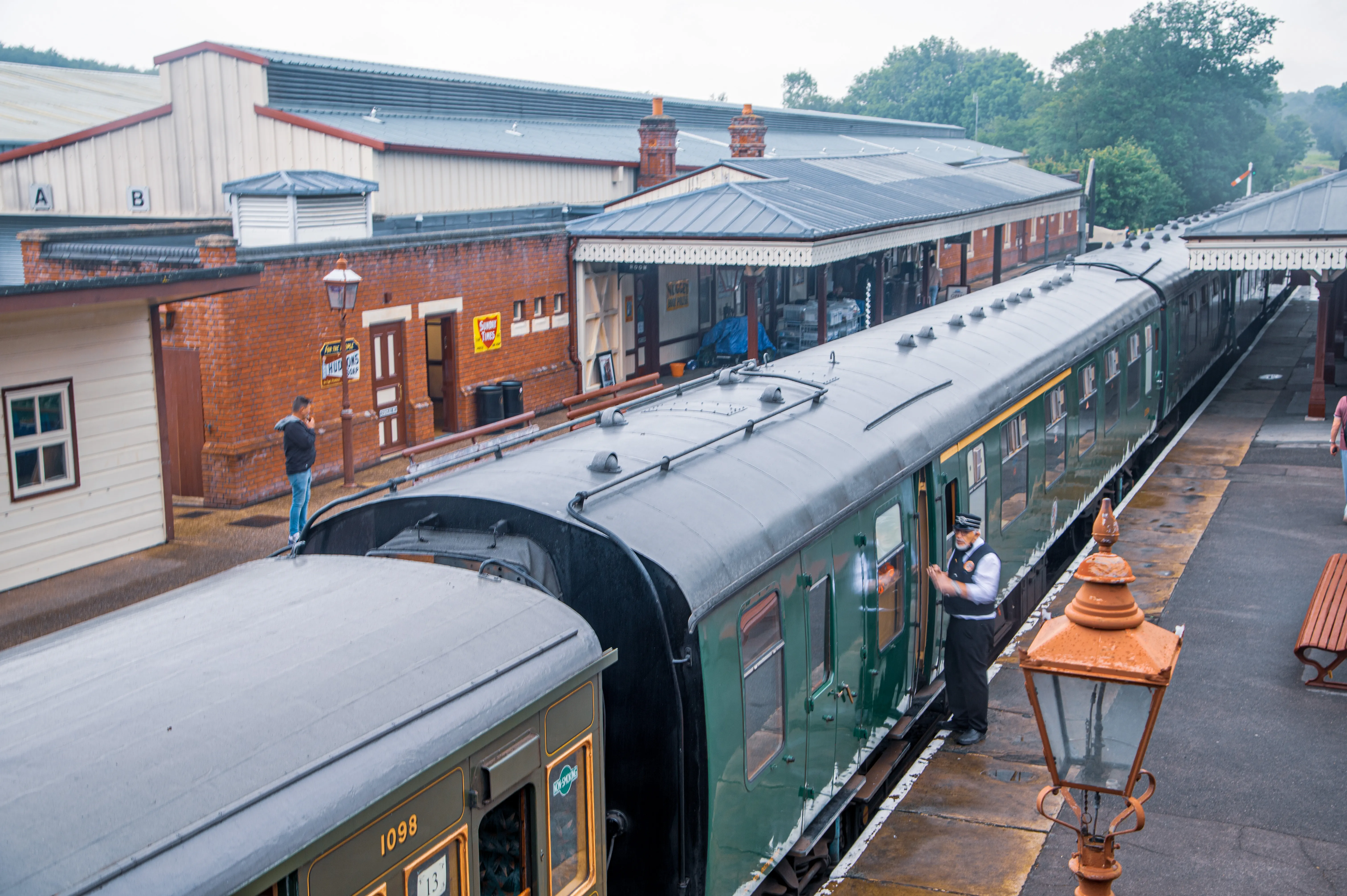 Green passenger train at a heritage railway station with uniformed staff and classic brick-and-timber buildings.