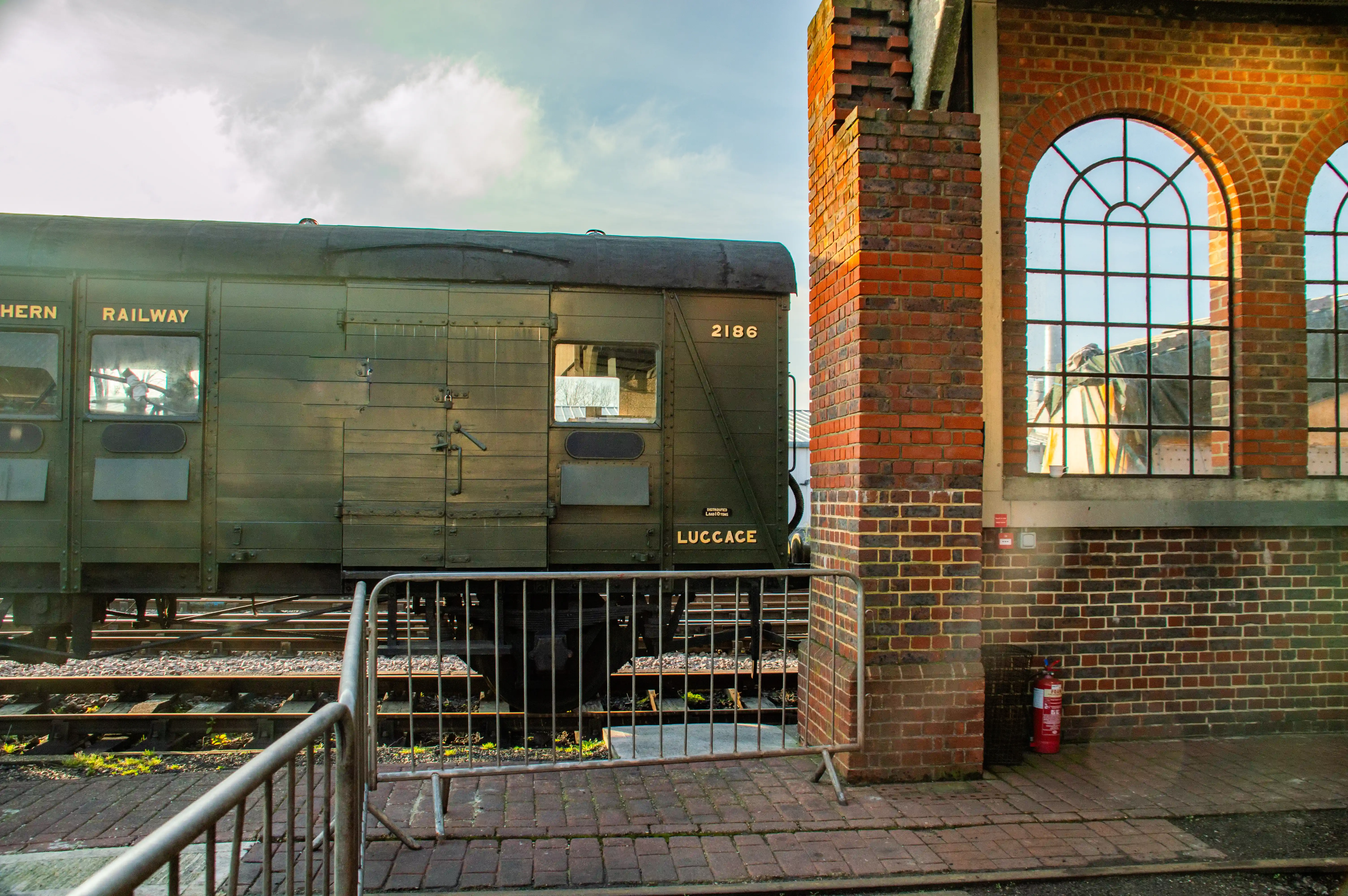 A vintage luggage train carriage with SOUTHERN RAILWAY and LUGGAGE visible, parked next to a red brick building with large arched windows reflecting the sky and some interior elements.