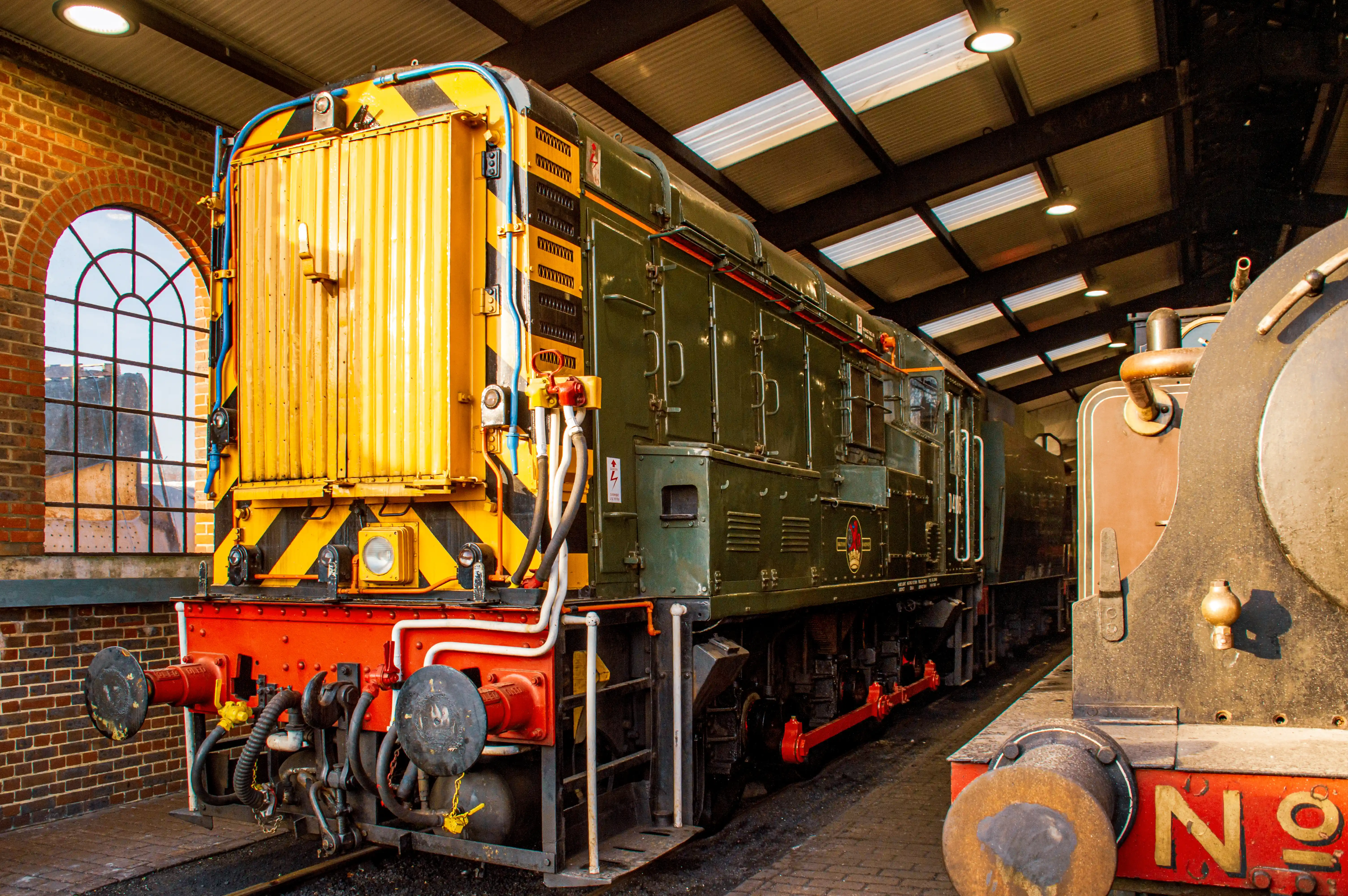 A close-up of the front of a dark green diesel locomotive with a bright yellow and black striped end, parked in a brick railway shed with large arched windows.